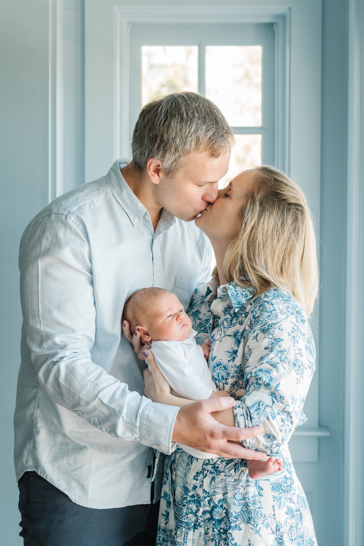 Parents kissing while holding newborn baby in front of window and blue wall - Northern Virginia Newborn Photographer