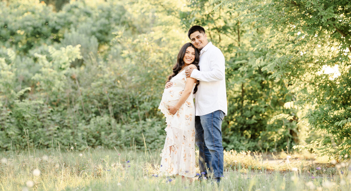 The picture is of a young couple capturing their maternity session portrait. The expecting couple stands in a lush green field with a forestry backdrop. The scene is beautifully captured by photographer Bri Sullivan in a light and airy style. The sunset adds a romantic touch to the portrait, highlighting the love and excitement shared between the couple.