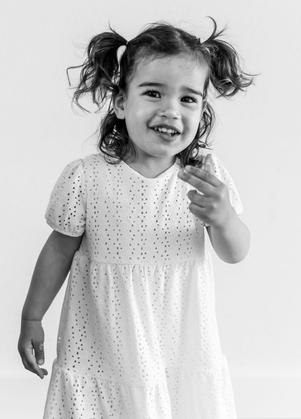 A candid, black and white portrait of a little girl with a mischievous smile. She has curly hair styled in two pigtails and is looking directly at the camera with her hand raised slightly. She is wearing a white dress with eyelet cutouts, and the background is a simple white.