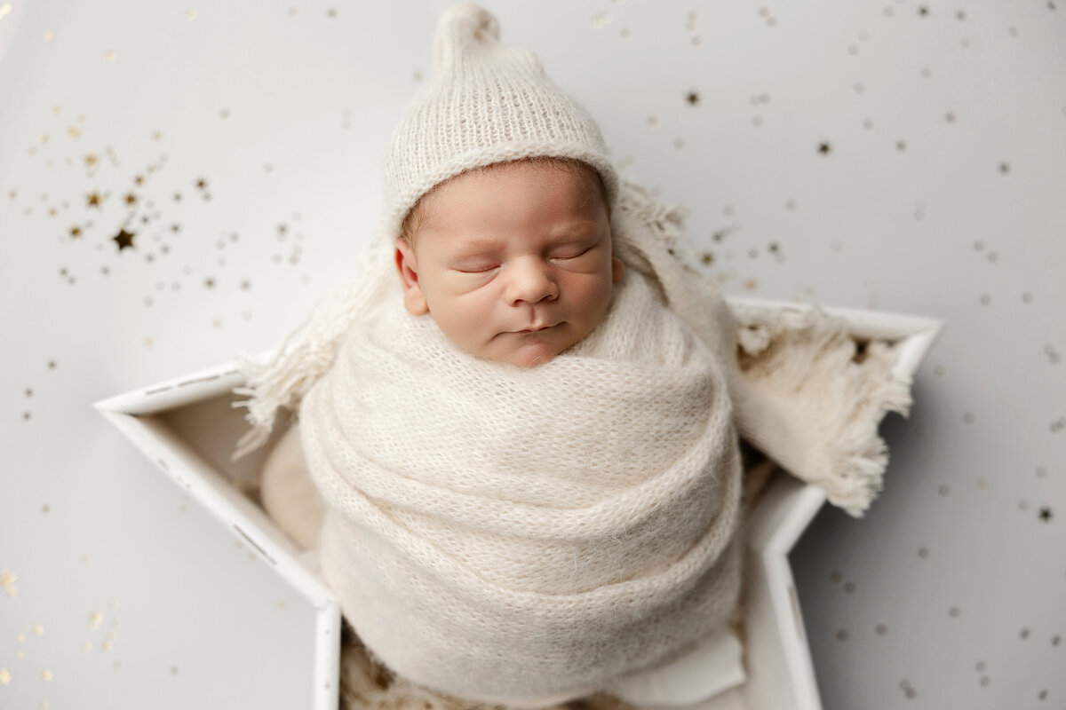 Newborn baby boy swaddled in soft cream knits with a matching bonnet, posed in a white star prop with scattered gold stars. Captured by Inland Empire newborn photographer Kaitlyn Dawn Photography.