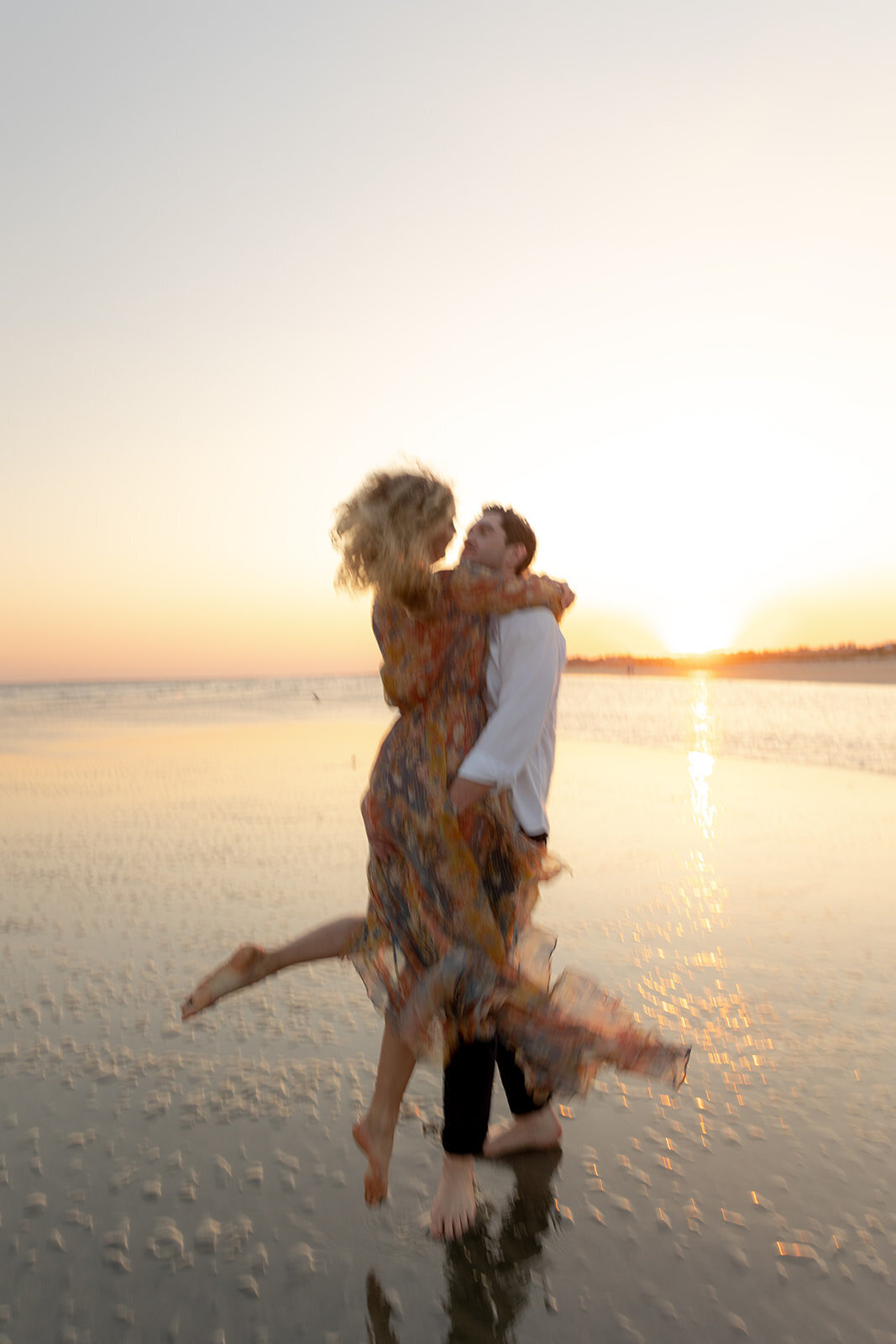 Charleston Beach Engagement Photo of man lifting up woman in colorful dress and twirls. Sun is setting at the horizon and reflecting on shallow water.