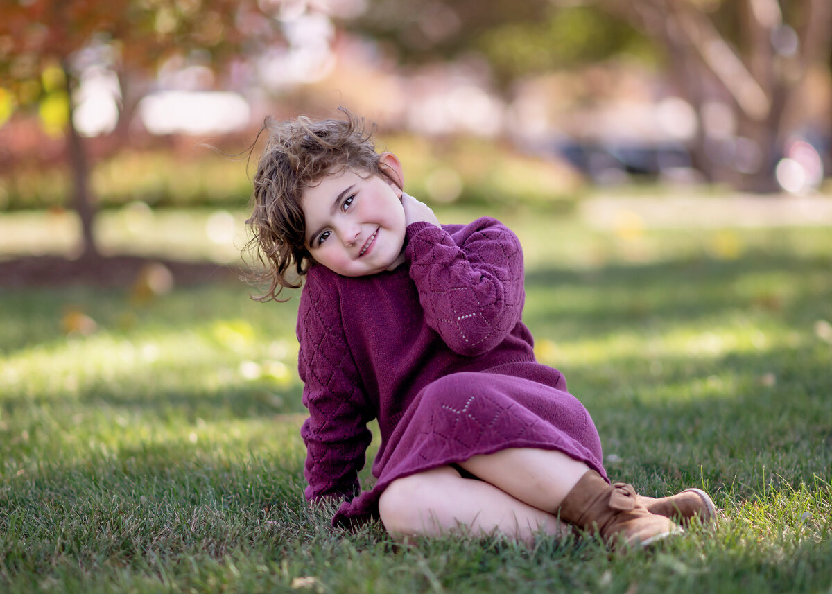 Child in a purple outfit plays with her hair as she sits on grass with autumn trees in the background