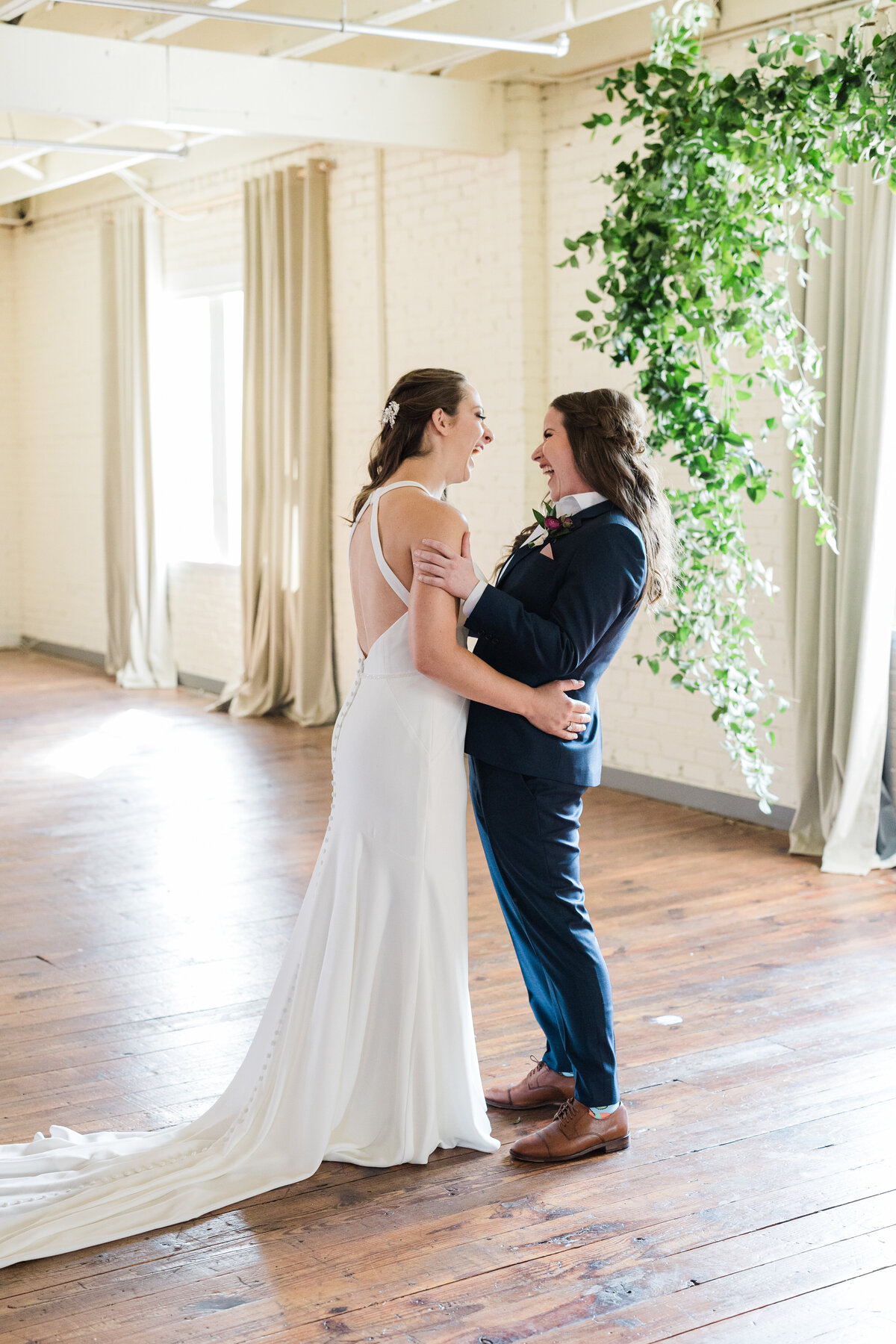 A candid shot of two brides during their first look before their wedding ceremony at the BRIK Venue in Fort Worth, Texas. They are both pulling each other close and laughing. The bride on the left is wearing a long, flowing, white dress, and the bride on the right is wearing a blue suit with brown dress shoes and a boutonniere.