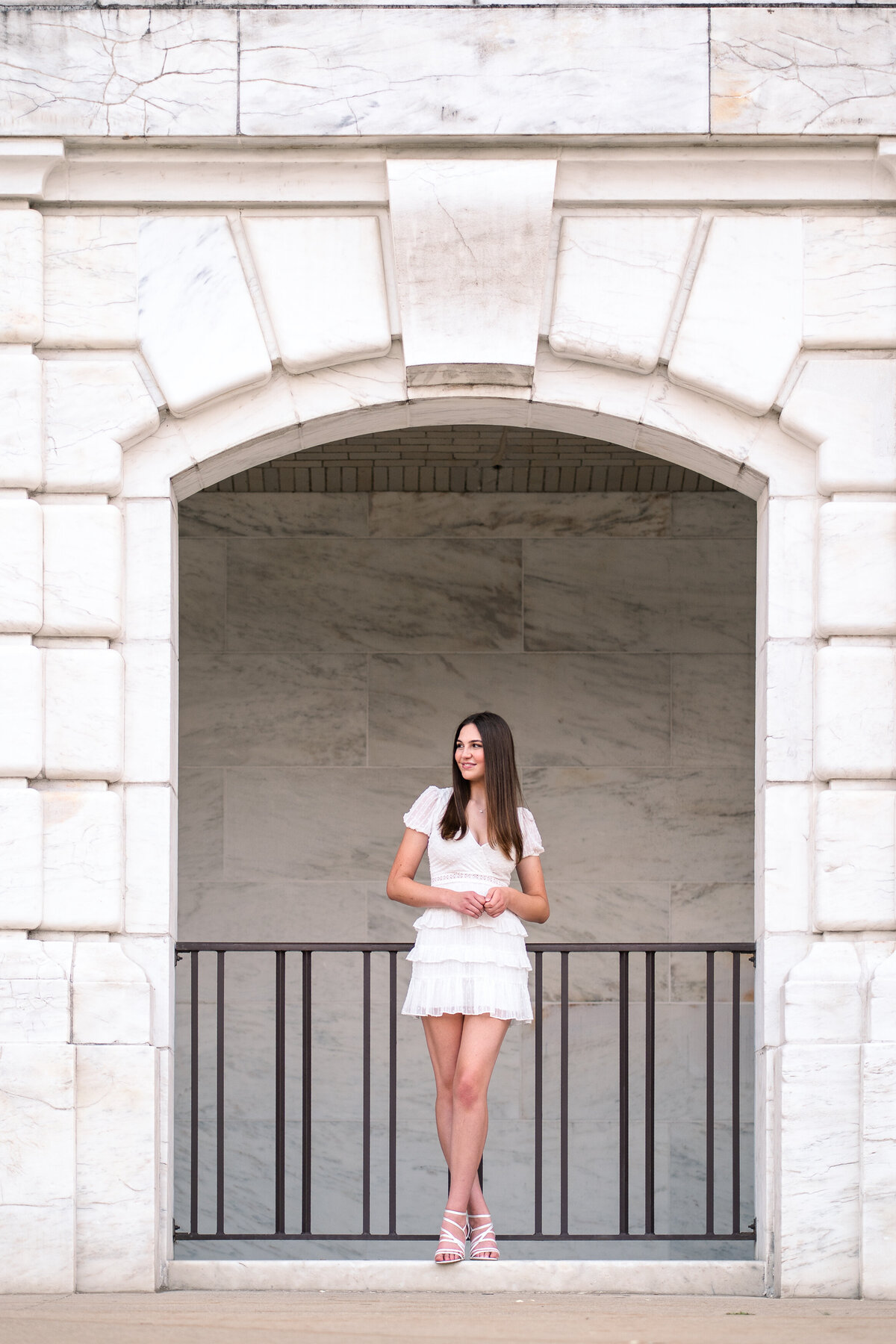 Teenage girl in a white dress standing agains metal gate under a stone archway looking off to the left as she is holding her hands.