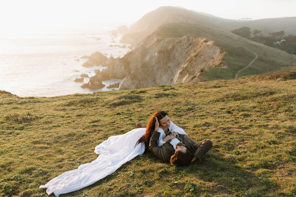 Couple in wedding attire laying down on grassy cliff overlooking the San Francisco Coast