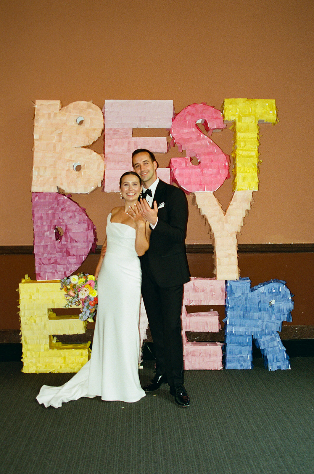 bride and groom standing in front of best day ever sign holding up their rings