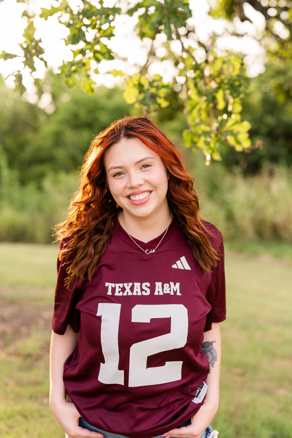 Texas A&M senior girl with hands in pockets and smiling while wearing Aggie maroon jersey and surrounded by nature at Leach Teaching Gardens
