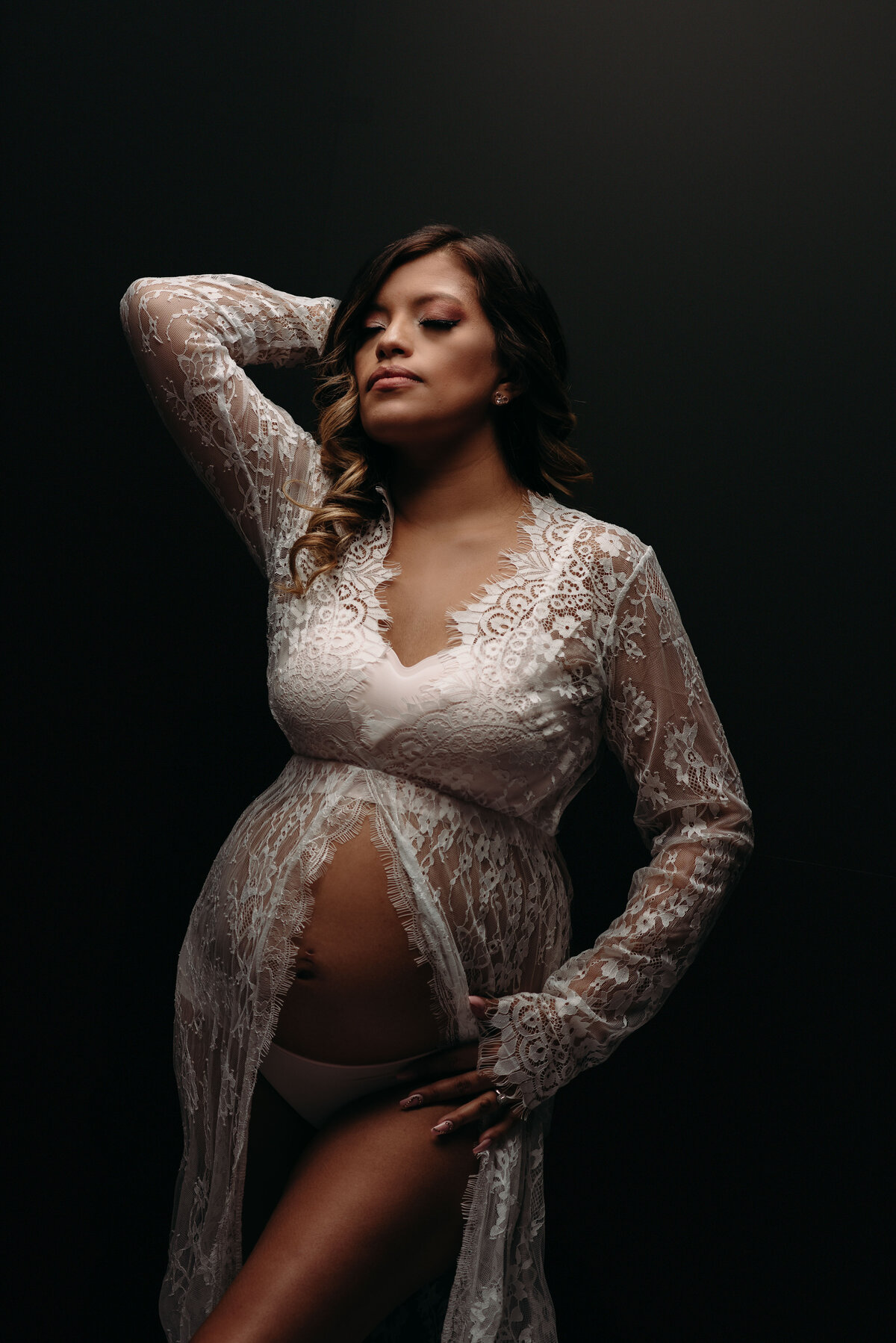 35 weeks pregnant woman wearing open lacy dress posing with one leg up and holding one hand on baby bump and other hand behind her head on black background