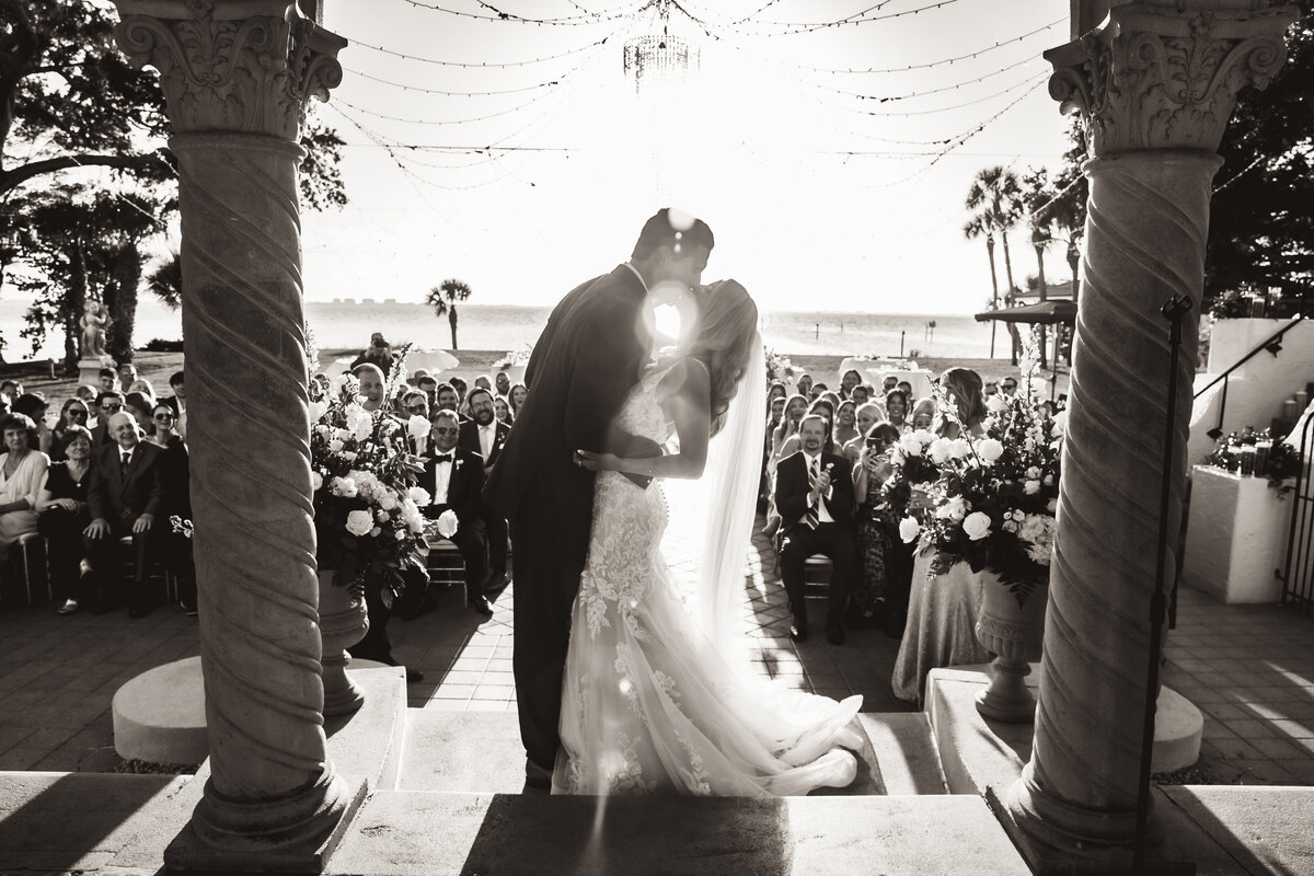 Shannon Lee, from Love Like Wild Photography, captures a newlywed couple sharing their first kiss in front of their guests, framed by elegant columns and sunlight.