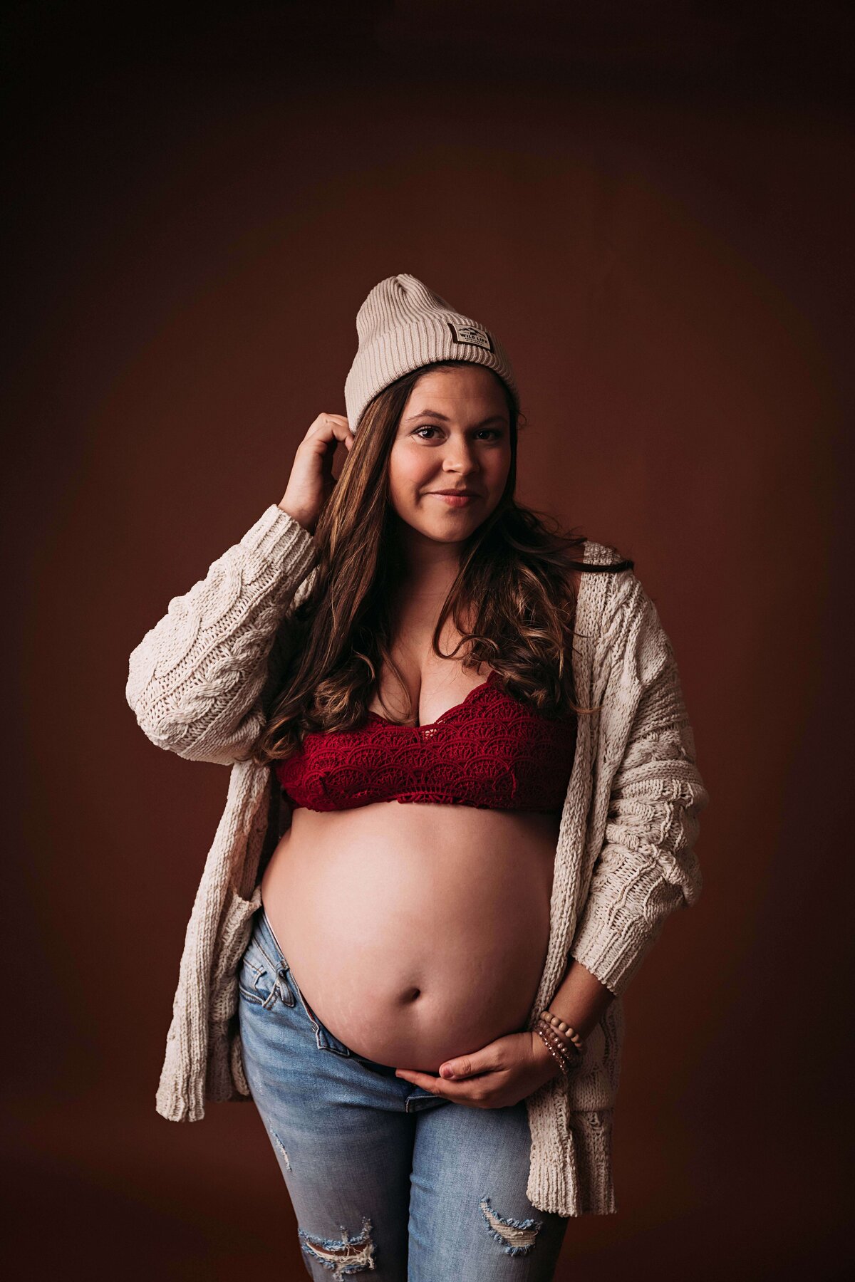 Close-up of a hipster mom with a beanie, red bra, and cardigan, softly smiling at the camera in this portrait taken by a Mobile, AL maternity photographer