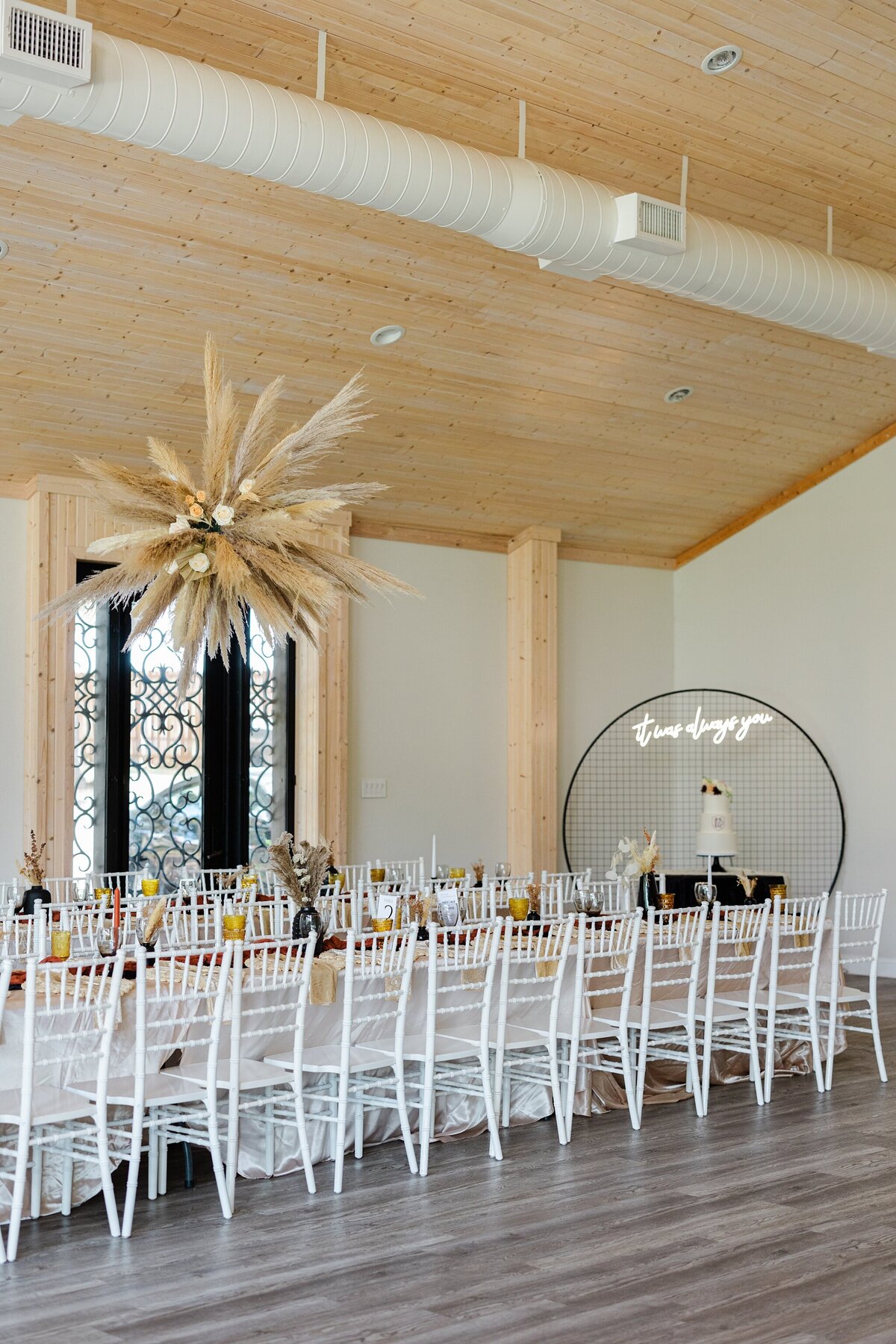 A detail shot of a reception room at a wedding reception in Dallas, Texas. A long, rectangular table can be see in the foreground. It's covered by a long, white tablecloth and is covered with place settings, napkins, cutlery, glasses, floral centerpieces and surrounded by white chairs. A similar table can be seen behind it. Above the tables is a large hanging floral decoration. Behind the table is the cake table with a three tiered wedding cake resting on it. Behind the cake hangs a neon sign reading "it was always you."