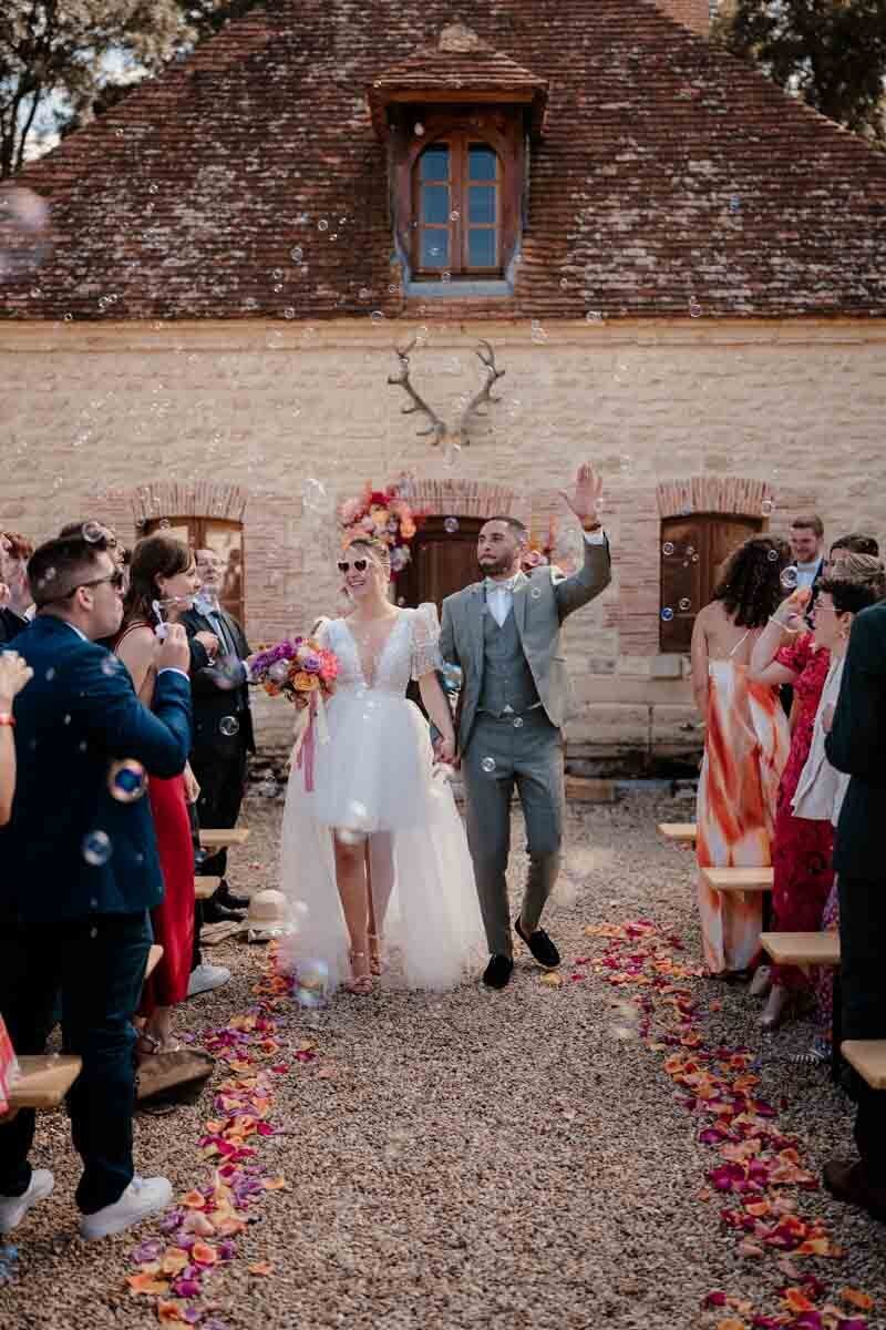 Sortie de cérémonie laïque joyeuse au Château de Vaux, avec les mariés en duo photo et vidéo, entourés de leurs invités soufflant des bulles.