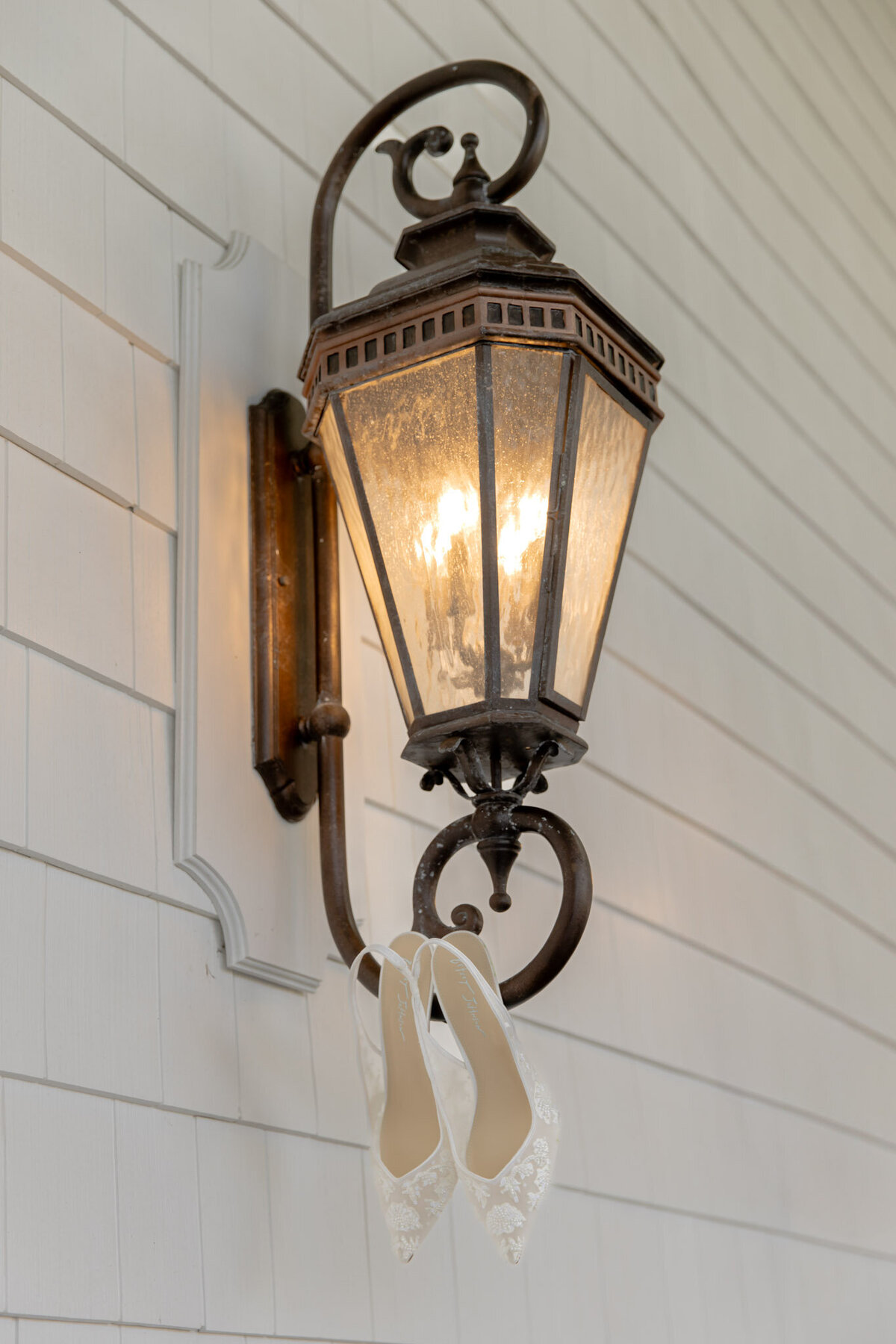 A light sconce on the outside of a white house. Hanging on it are the bride's wedding day shoes.