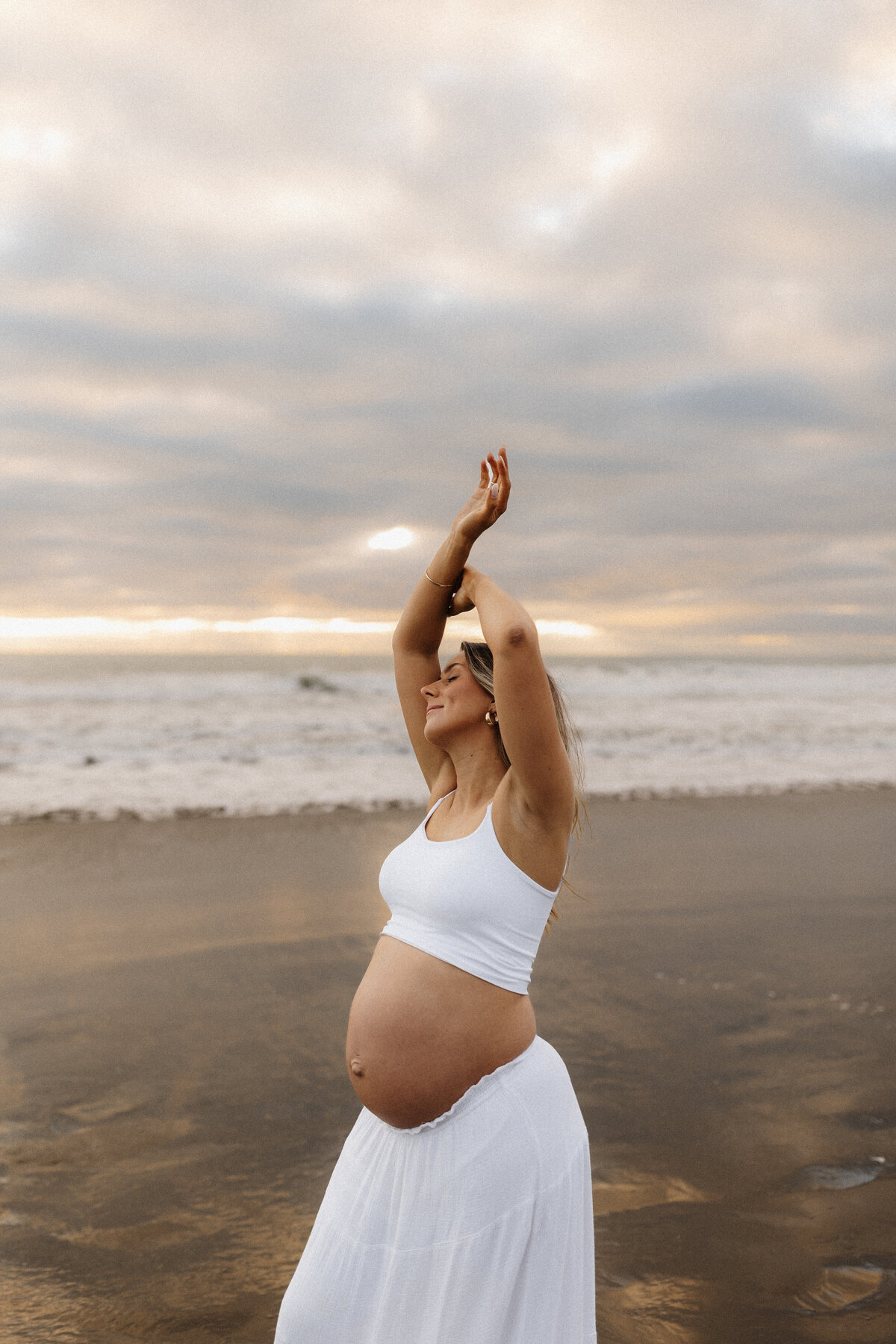 A mother to be wears a tank top and a white skirt and poses for the camera during a maternity photoshoot with christchurch photographer lucy jane photography