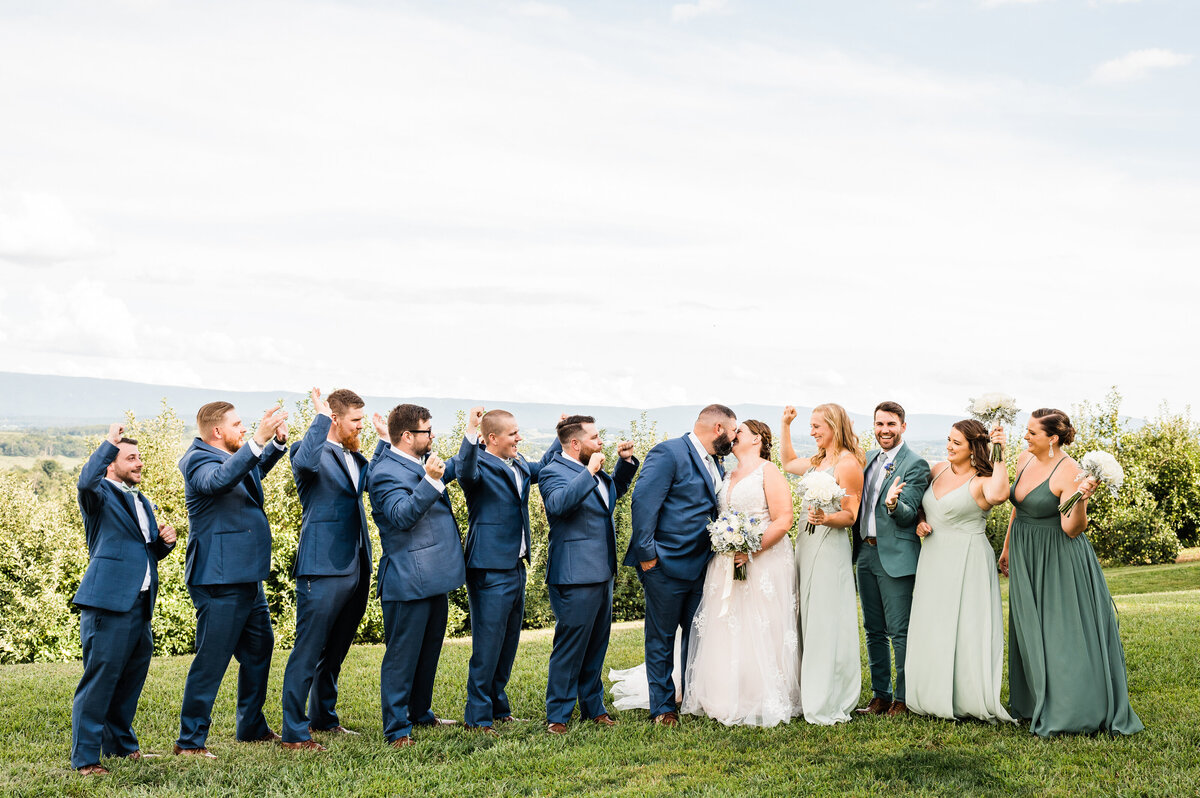 wedding portrait with bridal party surrounding the bride adn groom and cheering them on as the couple kisses romantically at showalter's orchard