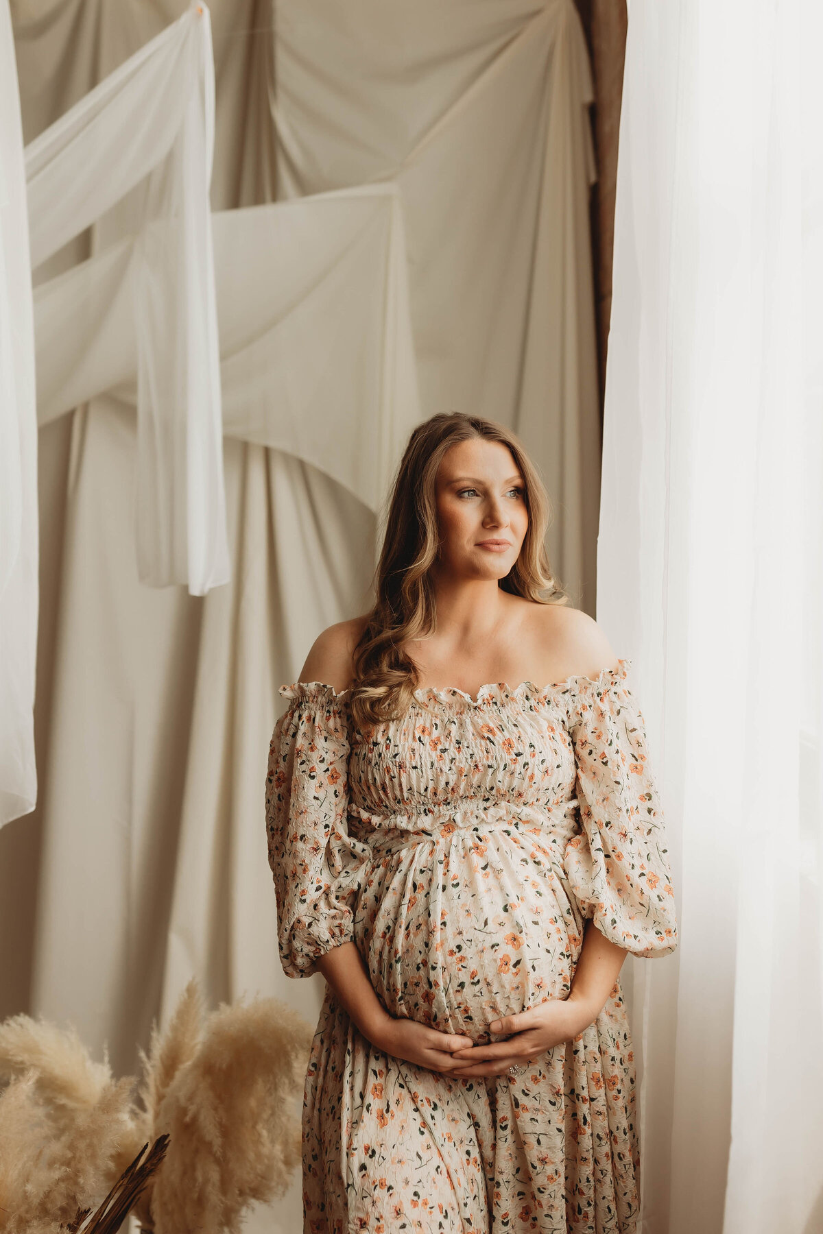 pregnant mother in a floral off the shoulder dress looking out the window in a dallas studio