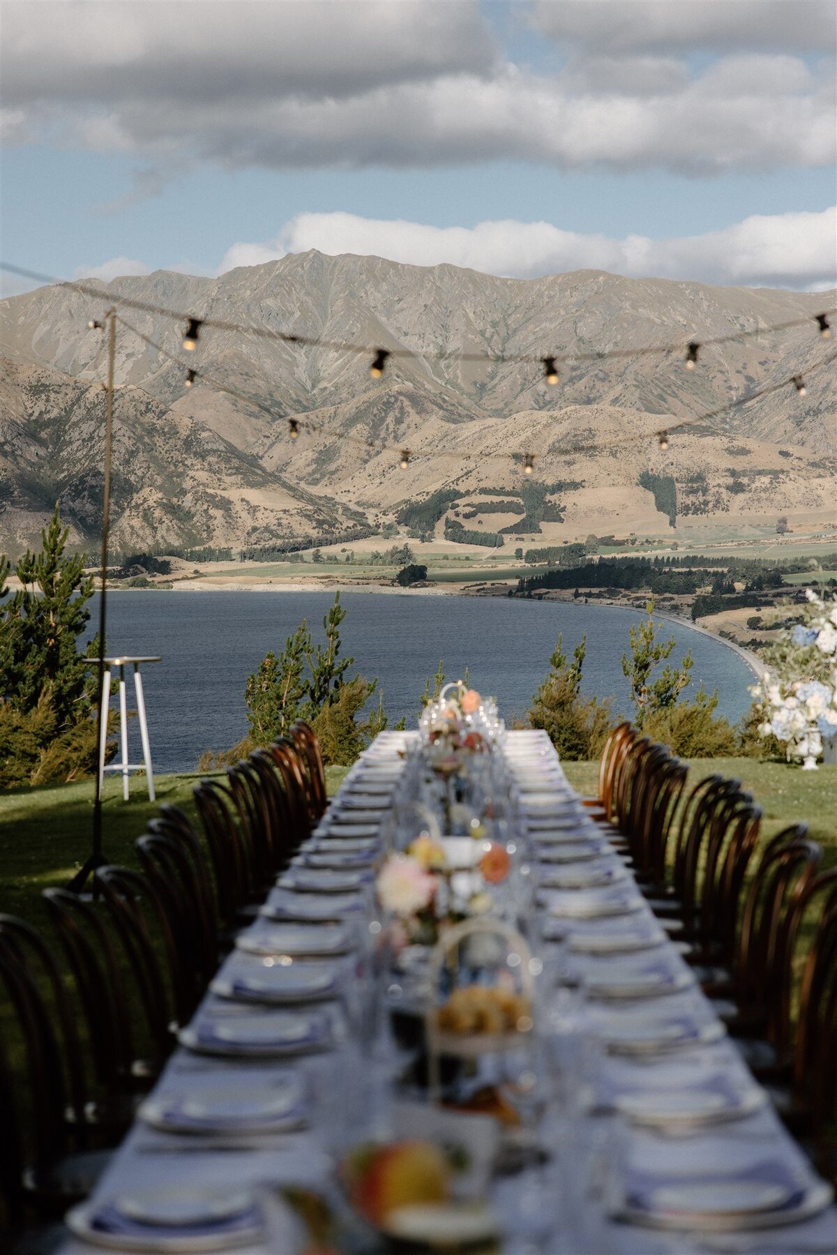 The long alfresco dining table stretches toward the curve of Lake Hāwea, framed by festoon lights and dramatic Central Otago peaks. This view alone is enough to stir goosebumps — truly a once-in-a-lifetime dinner setting.