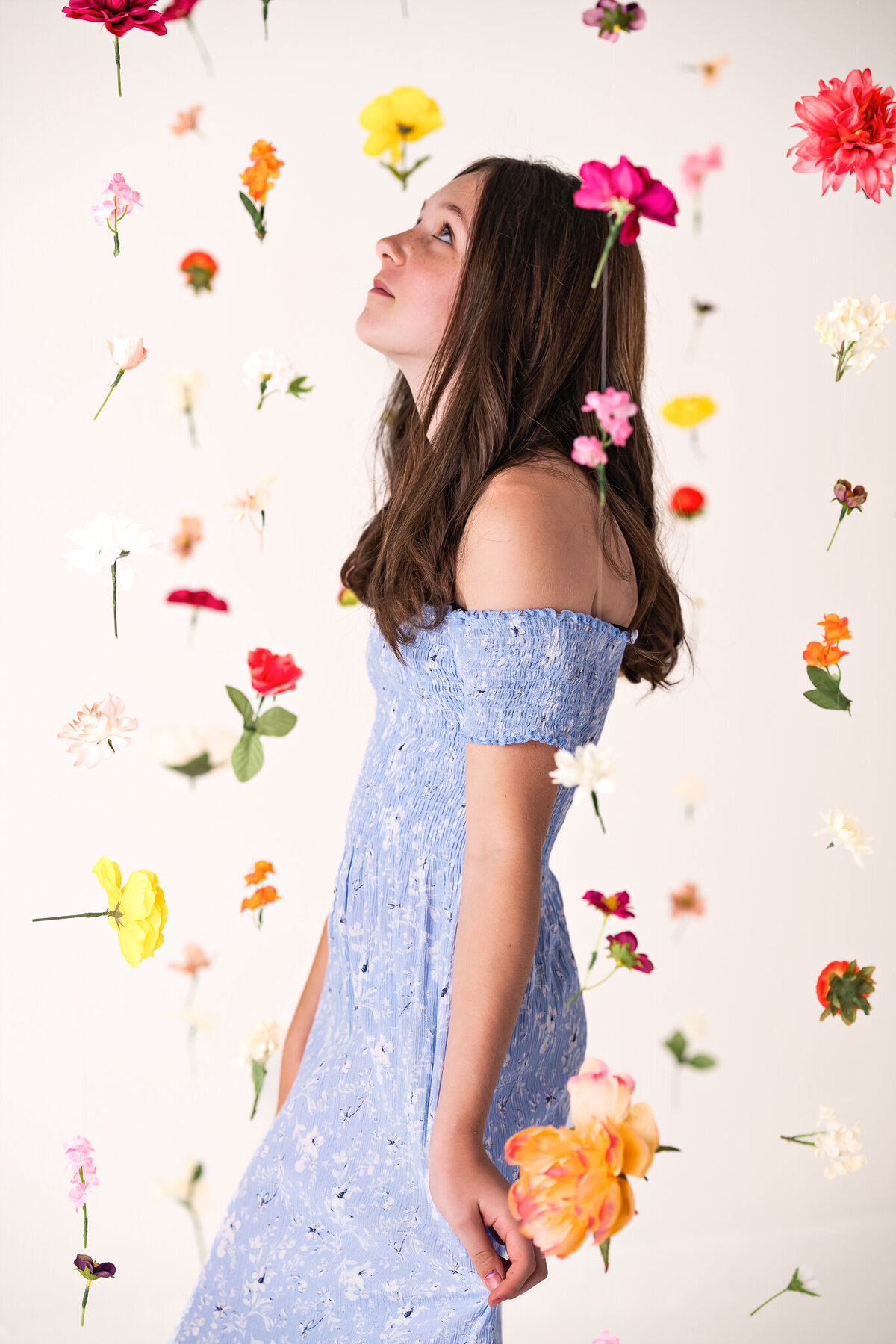 A teen girl in a blue dress is doing a photoshoot in a studio with hanging flowers.