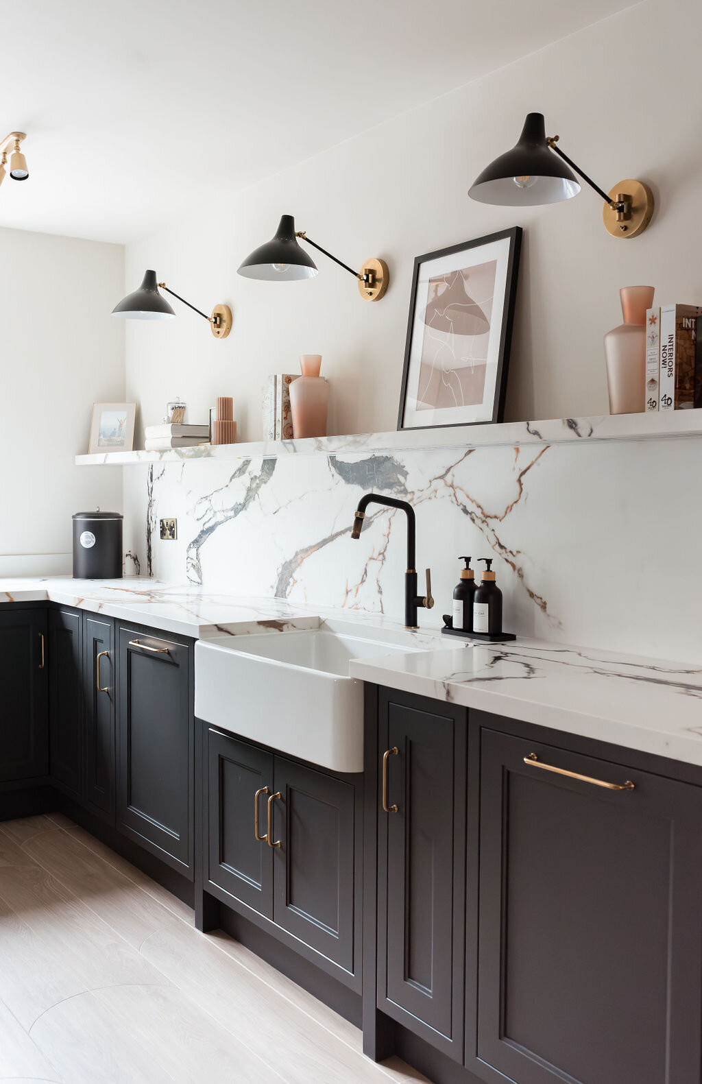 A modern kitchen with dark lower cabinets, a white marble backsplash with black and gold veining, a farmhouse sink, and minimalist open shelving holding books and decor. Two wall-mounted lights illuminate the space, which also features light wood flooring.