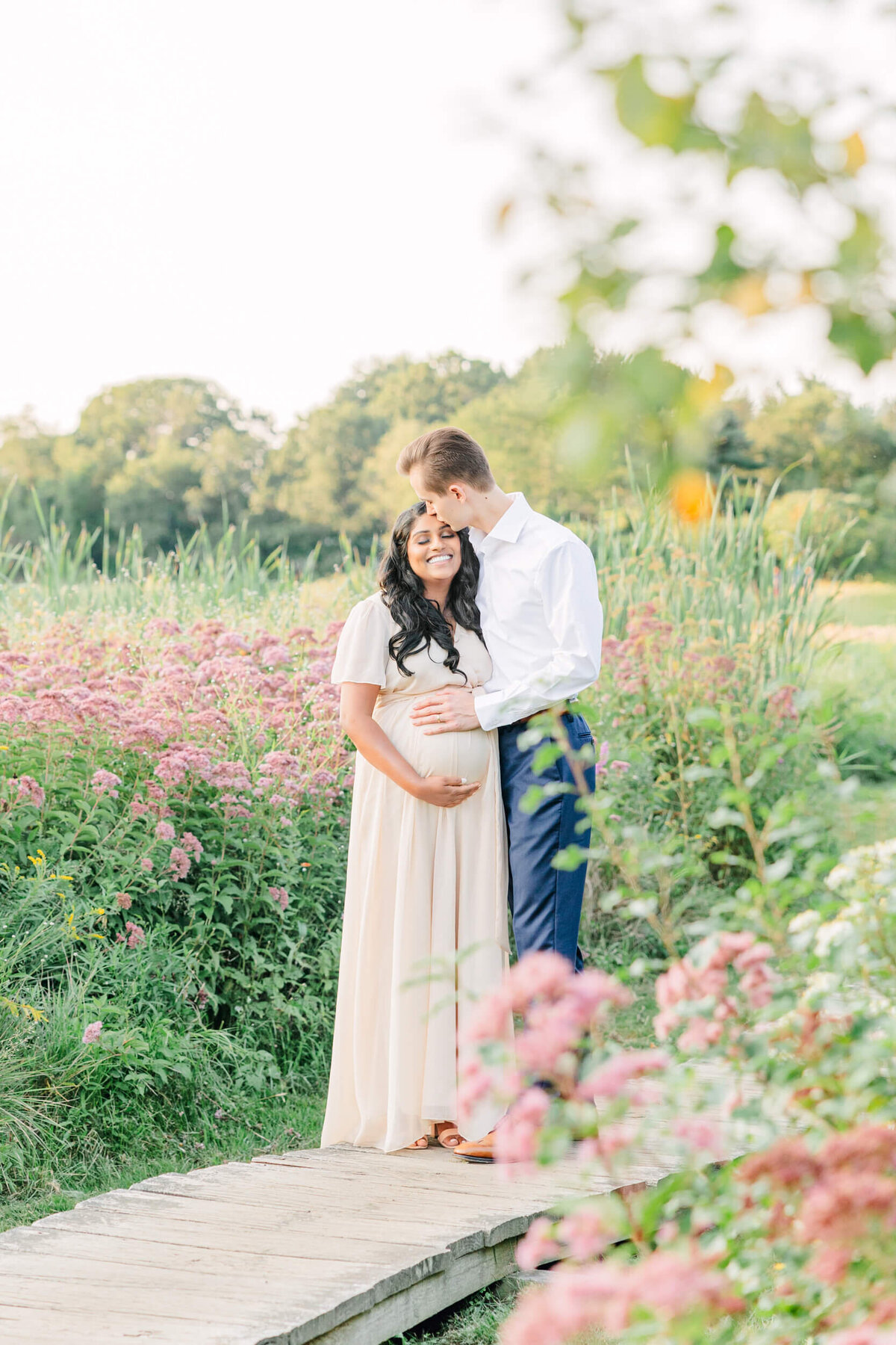 Tall man kisses the top of his smiling, pregnant wife in a field of pink wildflowers