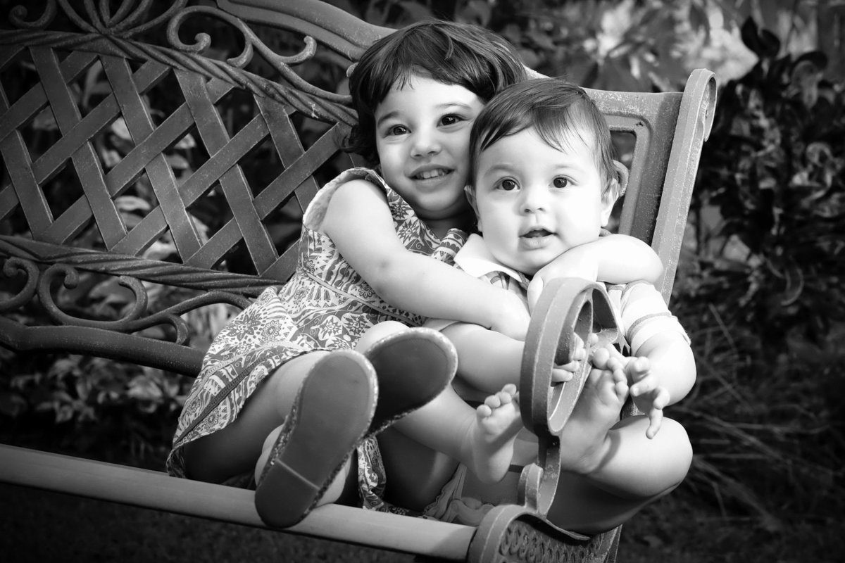 B+W shot of two toddlers hugging on an outdoor bench. Photo by Ross Photography, Trinidad, W.I..