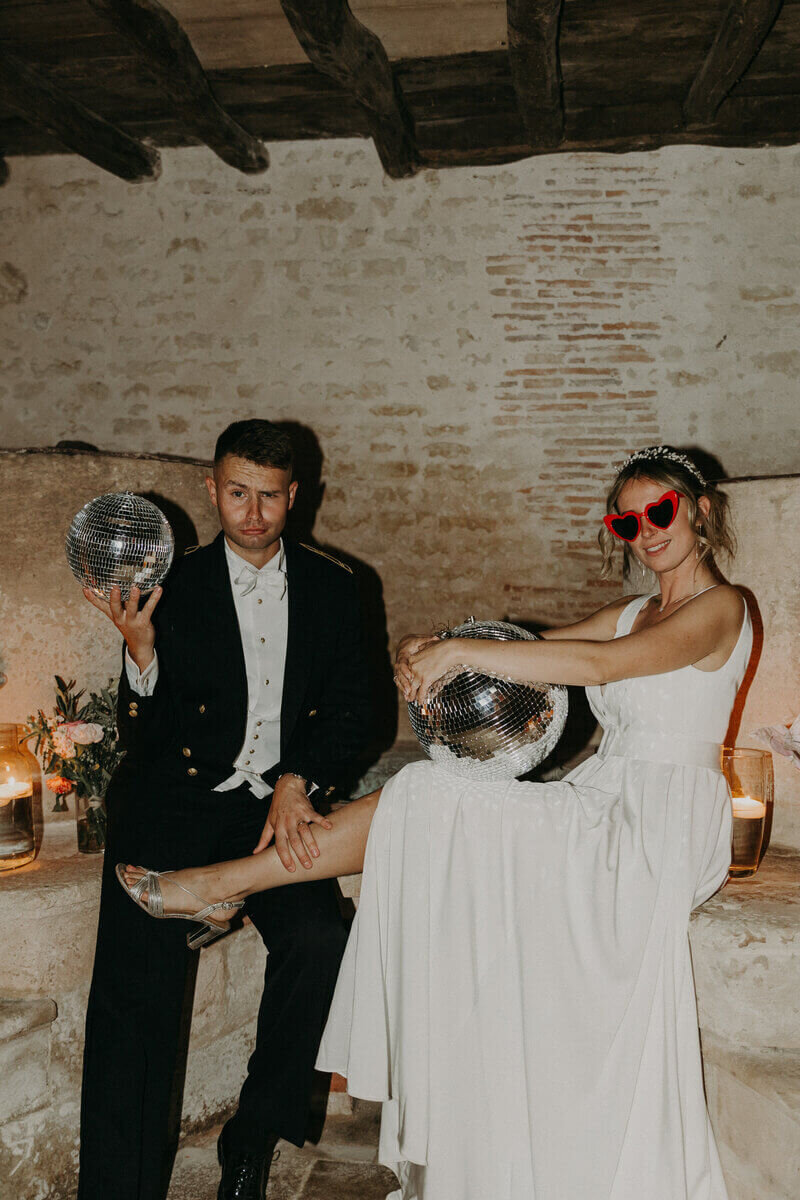 Marié en uniforme posant avec la mariée, lunettes rouges en forme de coeur sur le nez et tenant des boules à facette. Moment capturé par Laura Termeau photographie.