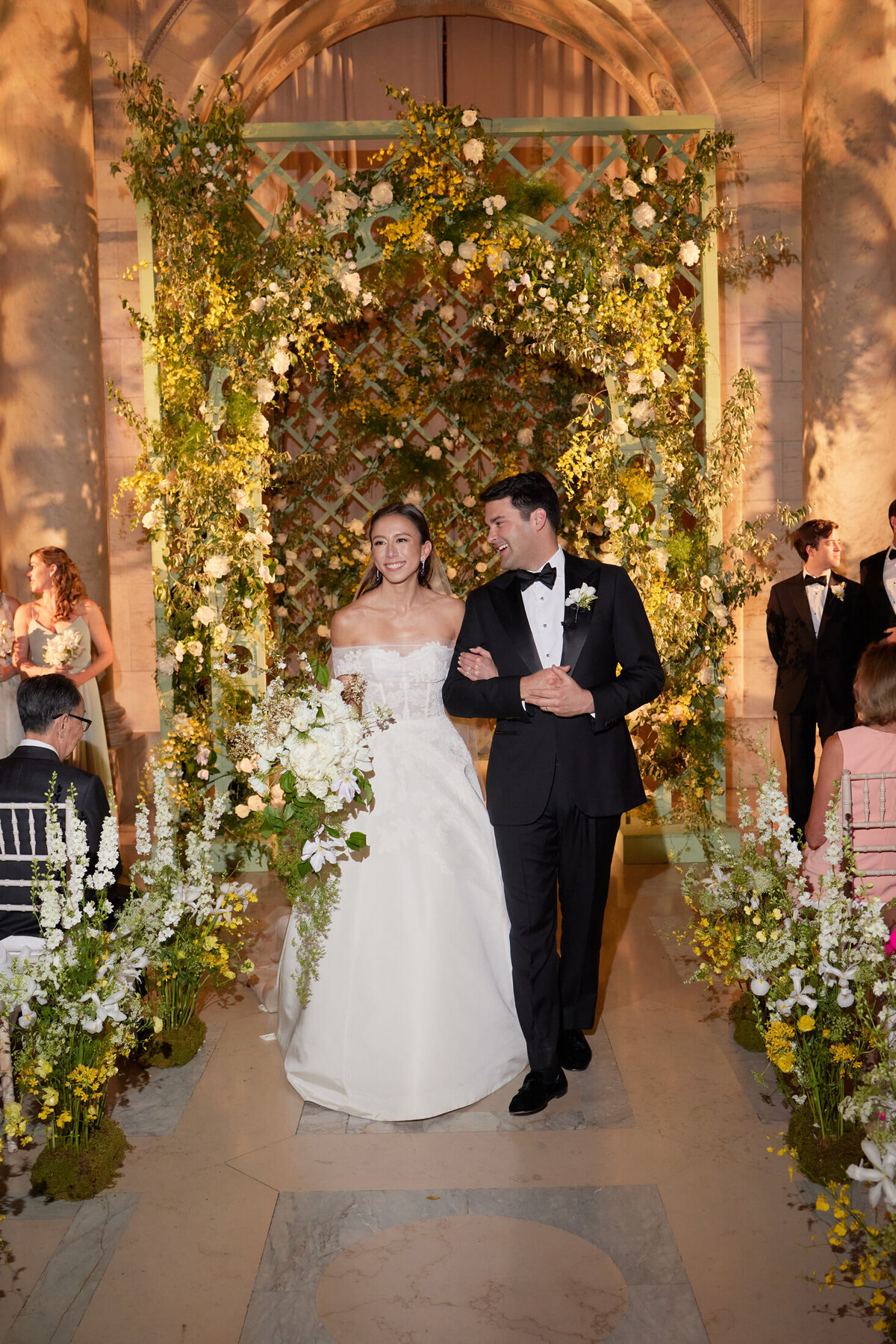 The photo was taken at New York City, The New York Public Library by photographer Shawn Connell. The bride and groom joyfully walk down the aisle after their wedding ceremony, surrounded by a beautiful floral archway and guests seated and observing their exit. In addition, you can add Aaron Navak Films as Videographers who captured this beautiful moment.