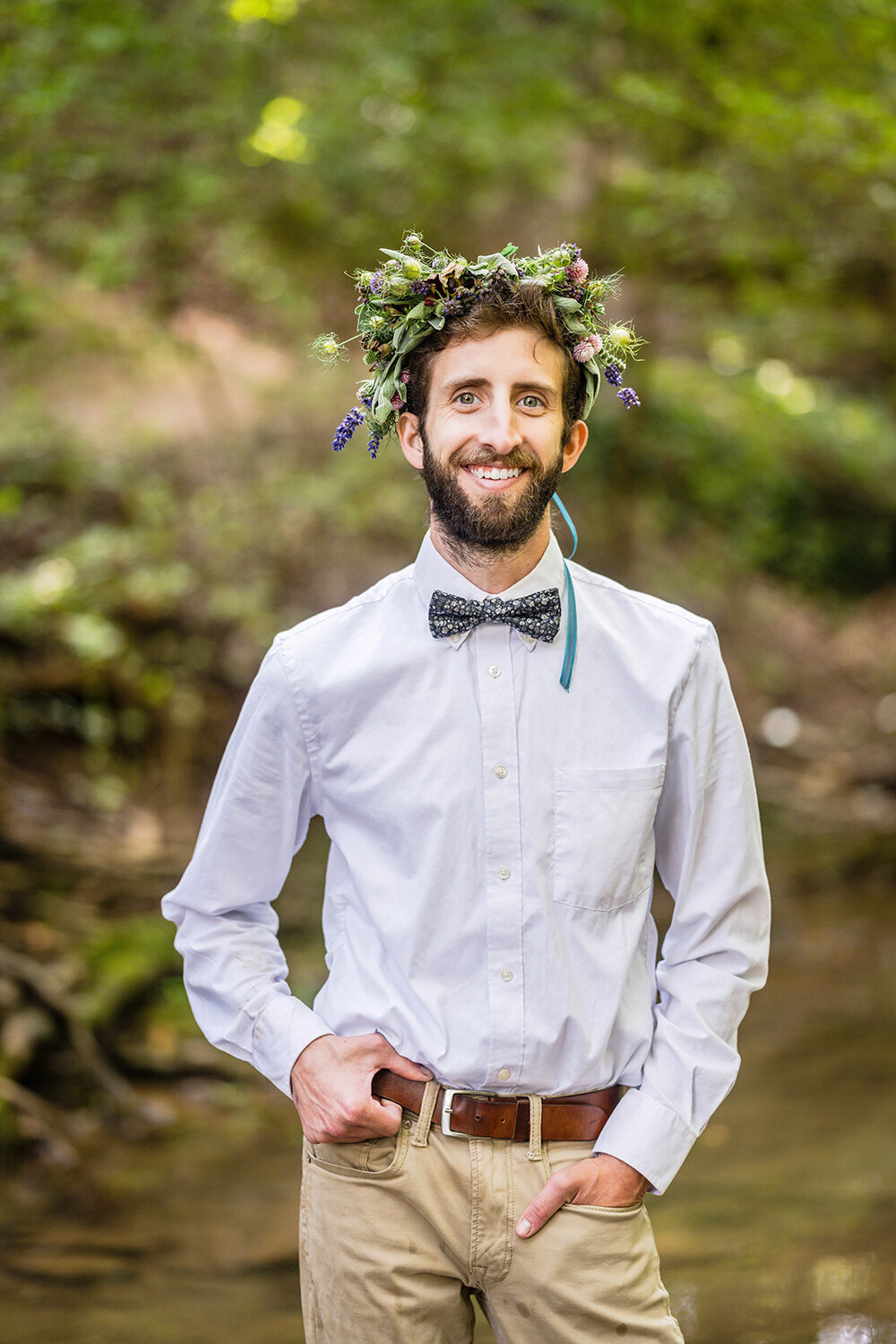 A groom smiles for a formal portrait on his elopement day. He is wearing a flower crown on his head, a simple white button up shirt, a bowtie, khakis, and a brown belt. He has one hand in his pocket while the other holds onto his belt.