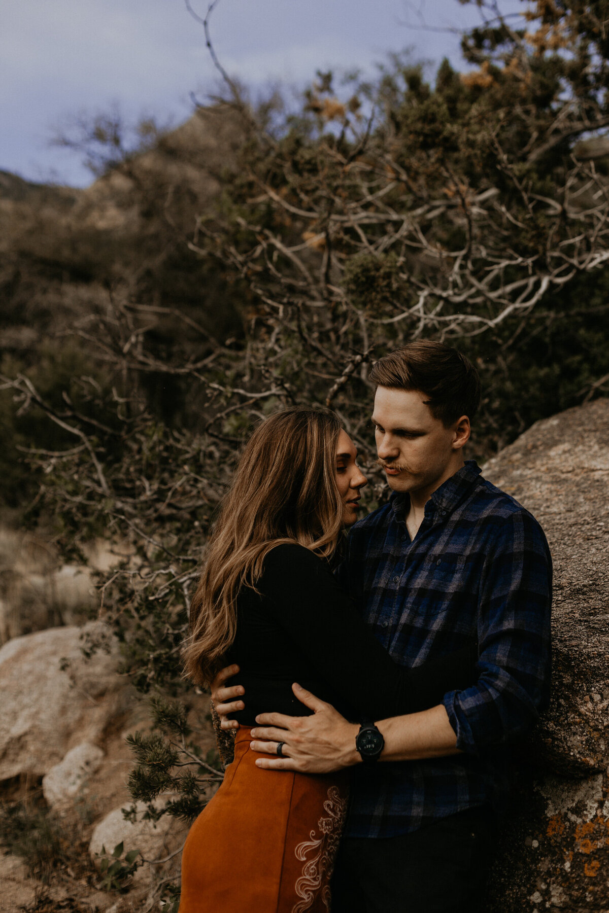 engaged couple leaning against a rock holding each other