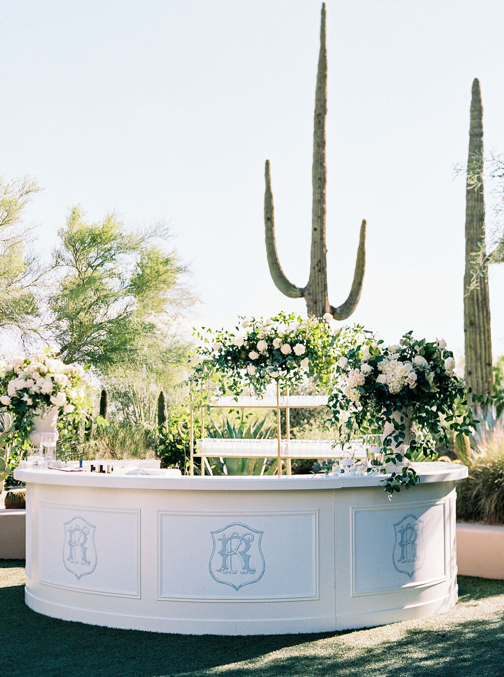 A sophisticated outdoor wedding bar adorned with fresh white florals and monogram details, creating the perfect luxury wedding reception experience under the open sky.