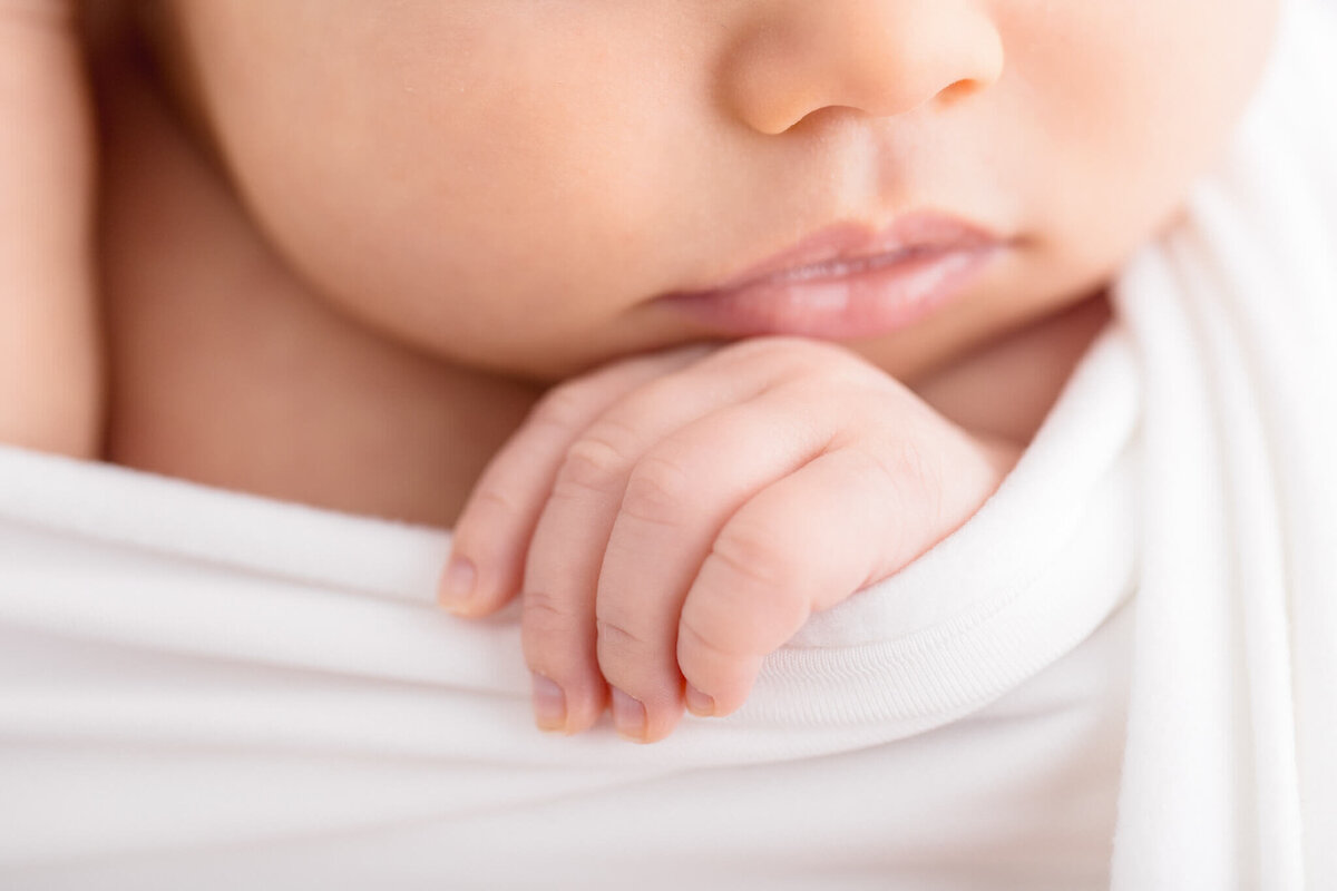 Close-up of a baby's hand gently resting on a soft white fabric. The image captures the babys lips and part of the face, conveying a sense of calm and tenderness.