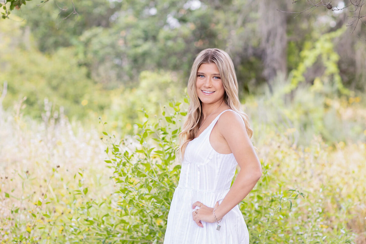 A woman with long hair stands in a lush green field, smiling, wearing a sleeveless white dress. The background is filled with blurred greenery and wildflowers.