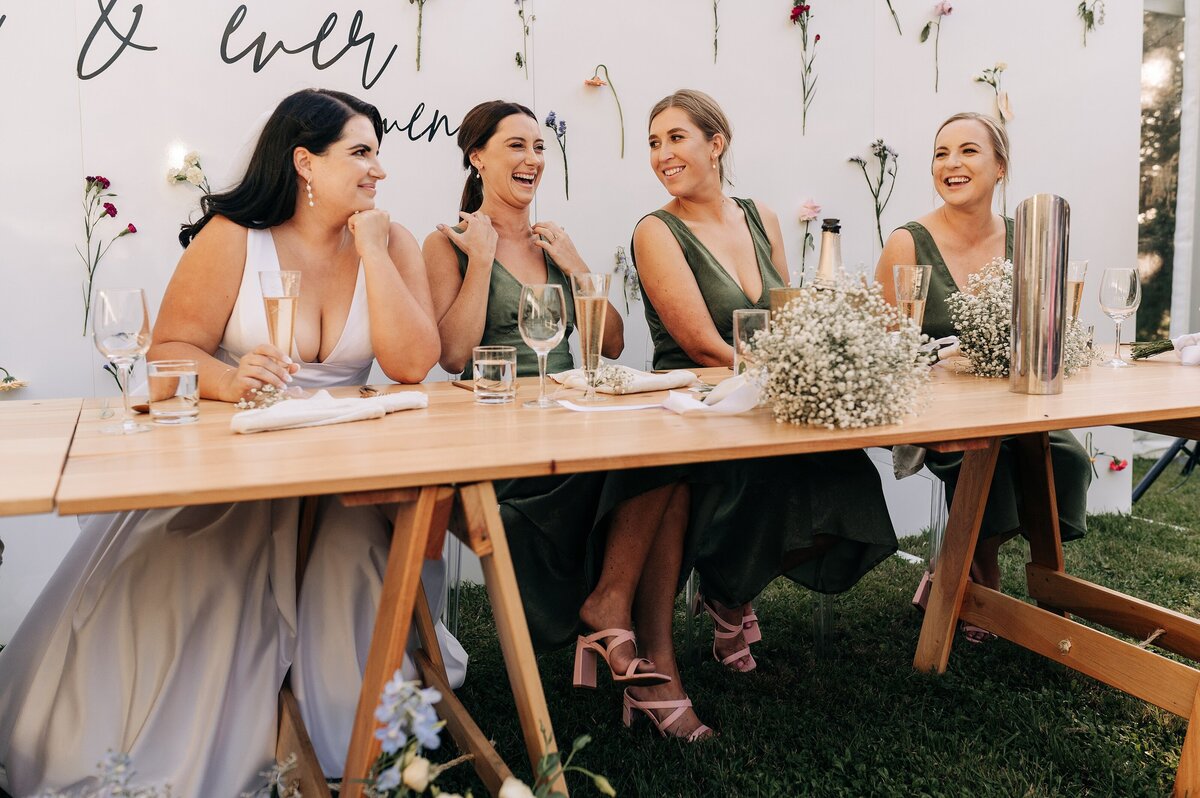 bride and bridesmaids in green dresses loula hire marquee and floral wall sitting at reception laughing loving ellies belly