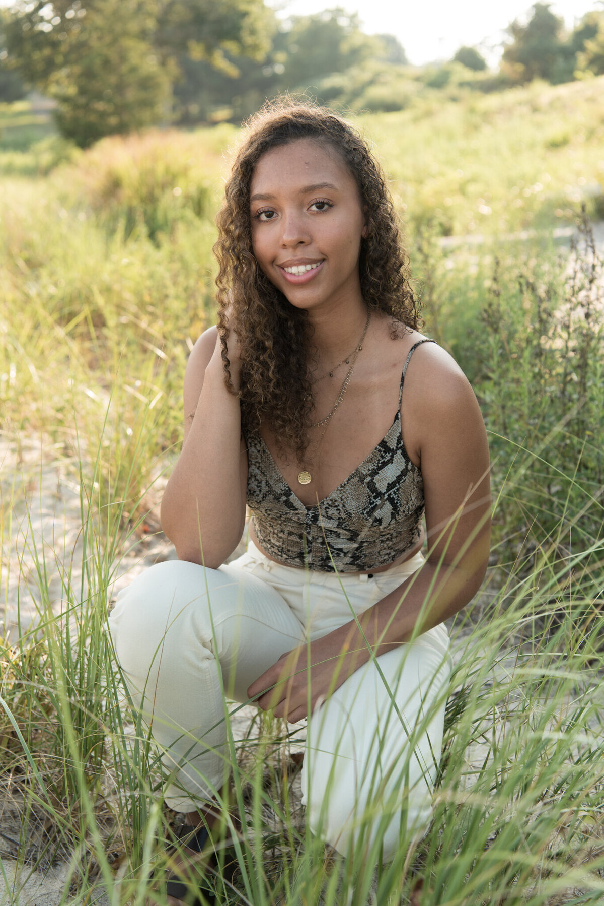 Teen girl squatting in dune grass with white pants and hand in her hair