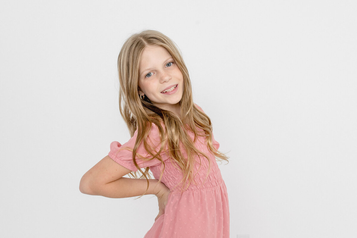 A pre-teen girl with long, blonde hair with her hands on her hips looking at the camera in a white studio in Tustin.