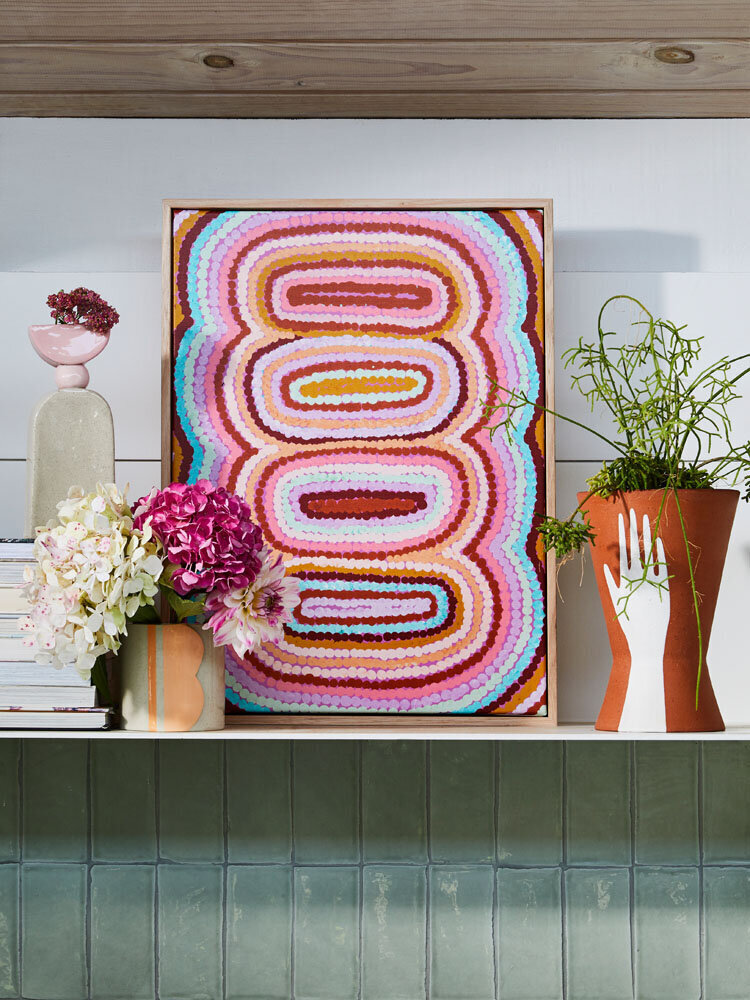 Close-up of a shelf with bright Aboriginal dot painting, flowers, and sculptural vases.