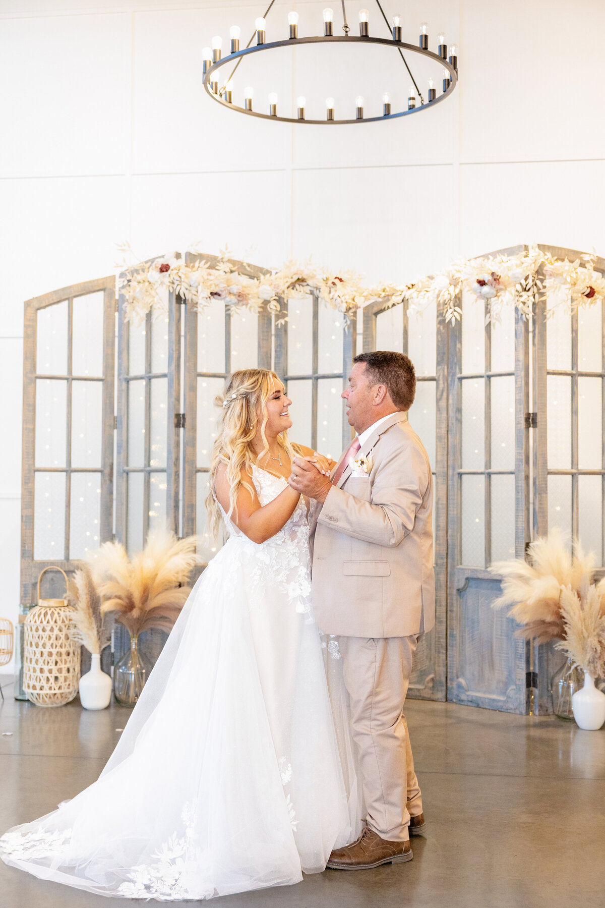 The bride and her father share a heartfelt dance, smiling warmly in an elegantly decorated reception space.