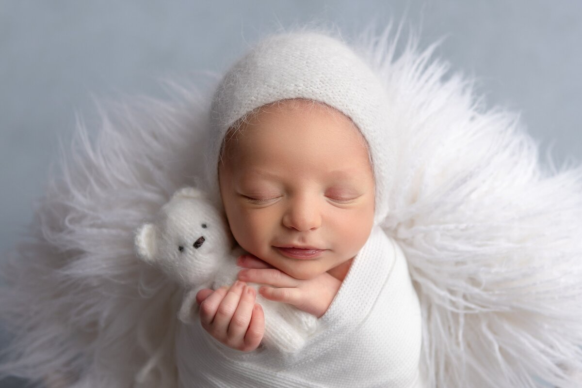 Newborn baby boy holds a teddy bear in white set up at a Wellington newborn photography studio.