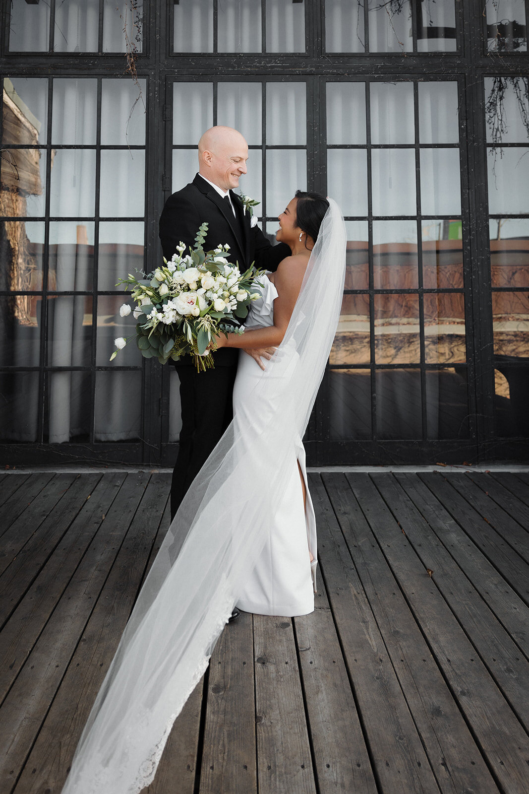 couple holds each other on wooden deck and brides veil blows in the wind