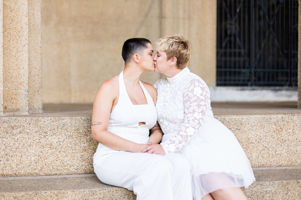 Gay women kissing wearing white outfits sitting on a step at LGTBQ wedding in Centennial Park, Nashville,TN