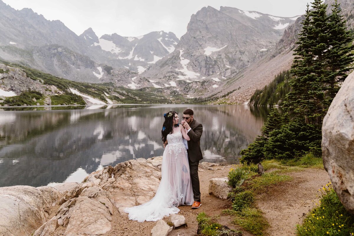 Bride and groom standing on a rocky shoreline during their Lake Isabelle, Colorado elopement, with calm alpine water and dramatic snow-capped peaks in the background.