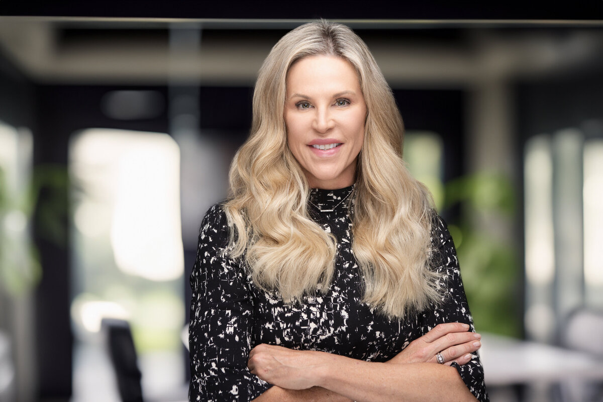 Professional headshot of a middle-aged Caucasian woman with long blonde hair, captured in Cincinnati for personal branding. She is wearing a stylish black and white patterned top, standing confidently with her arms crossed. This headshot showcases her approachable and professional demeanor, perfect for updating LinkedIn profiles and personal branding materials. Optimize your Cincinnati headshots with expert photography services for a polished, professional image.