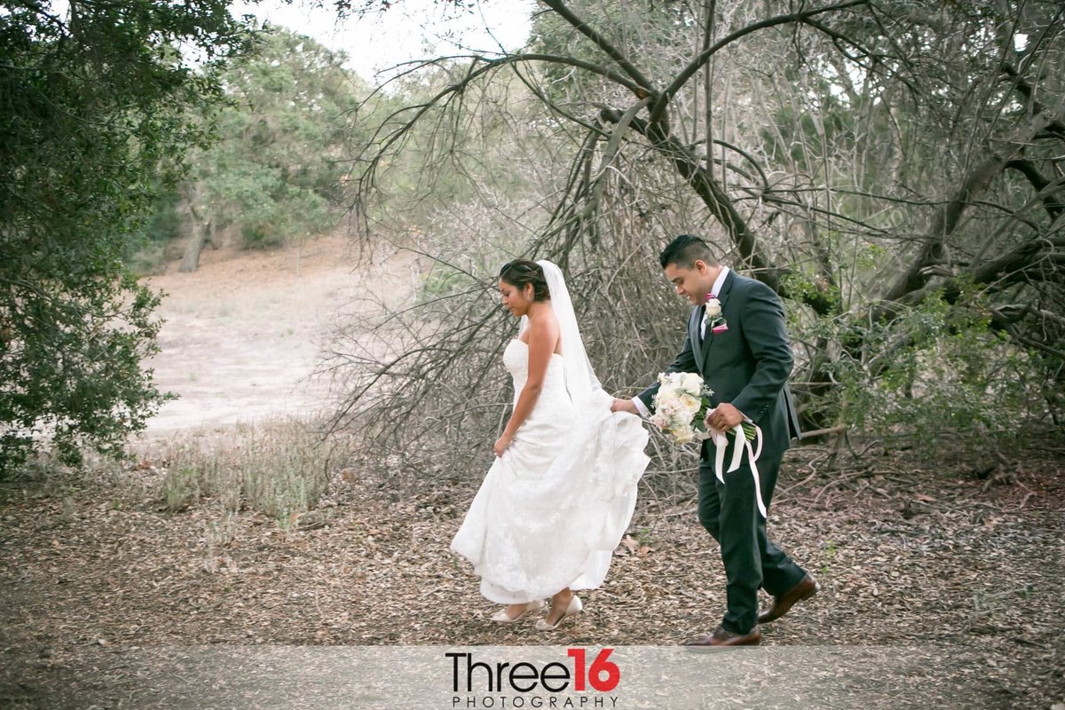 Newly married couple go for a walk in the woods as Groom carries her dress train