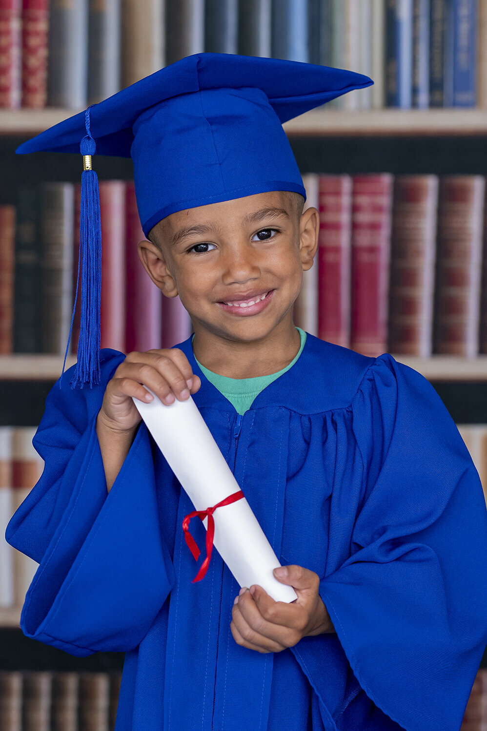 Boy holding his graduation certificate stands infront of a book case backdrop in his cap and gown smiling.