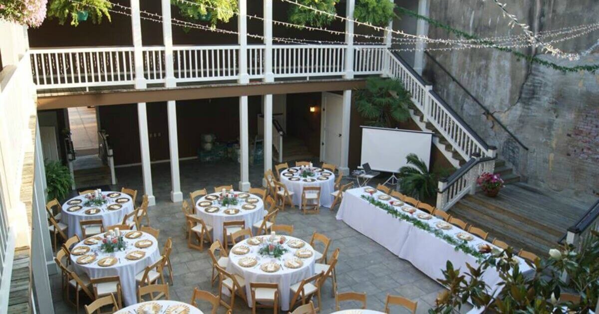 A bright courtyard set up for an event features round tables with light blue tablecloths, wooden chairs, potted plants, and string lights hanging above. The photo is looking down from above.