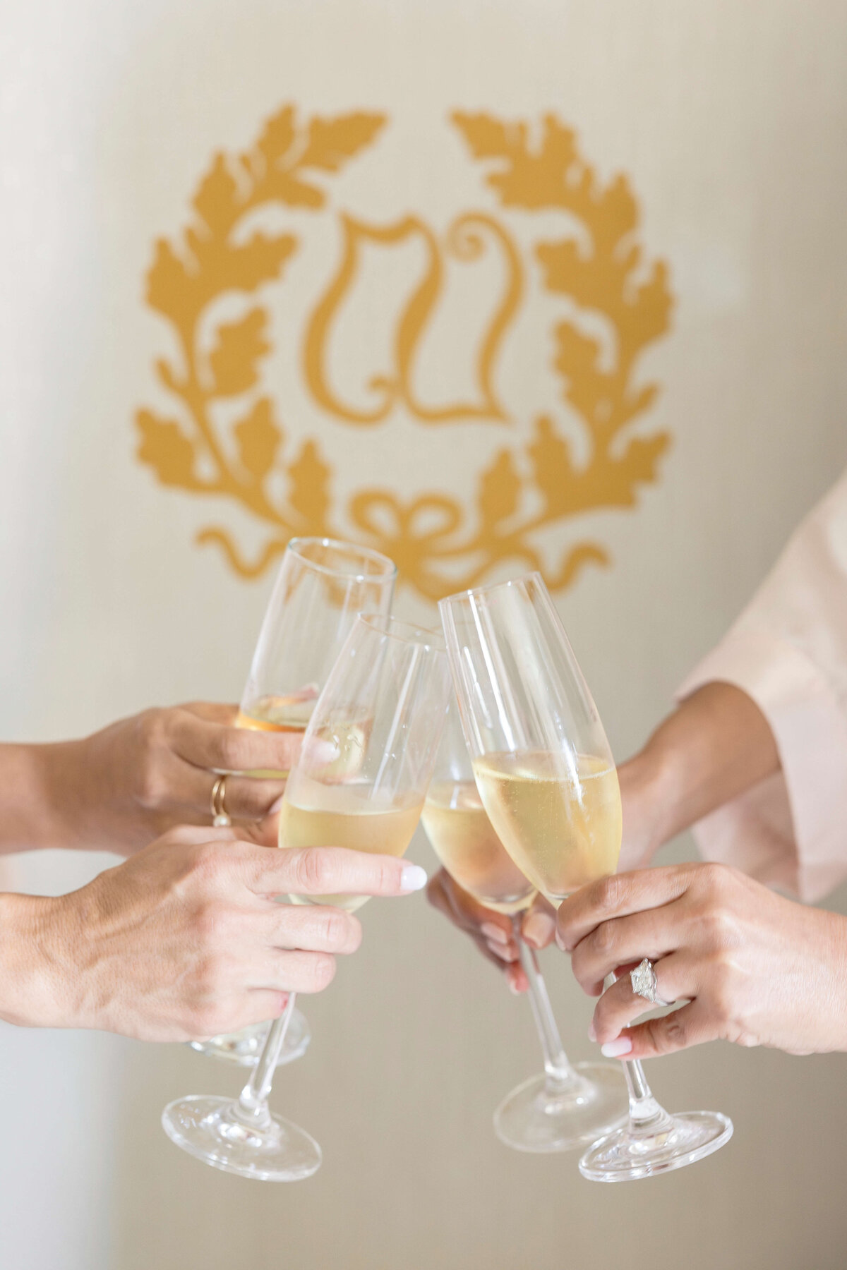 Four hands hold champagne flutes in a celebratory toast against a backdrop with a gold decorative emblem.