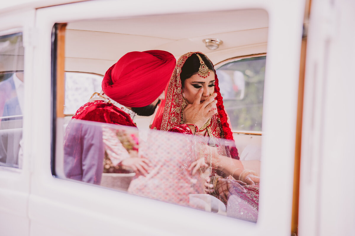 sikh groom stopping the tears from falling from the bride's face inside the car after their wedding ceremony during the doli vidai