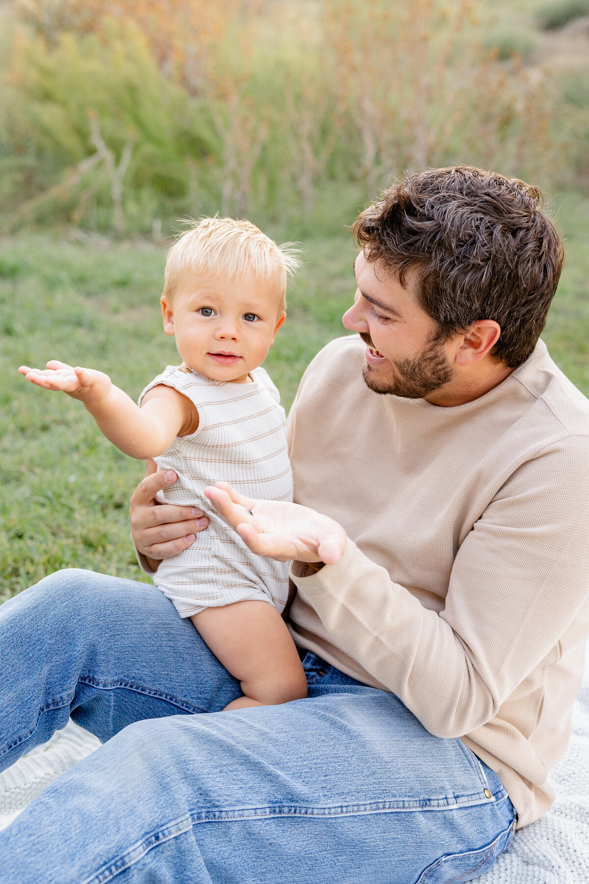 A father holds his baby on his lap, playfully gesturing as the baby mimics him with curiosity and delight. A touching father-son moment captured in Eagle Mountain, Utah.
