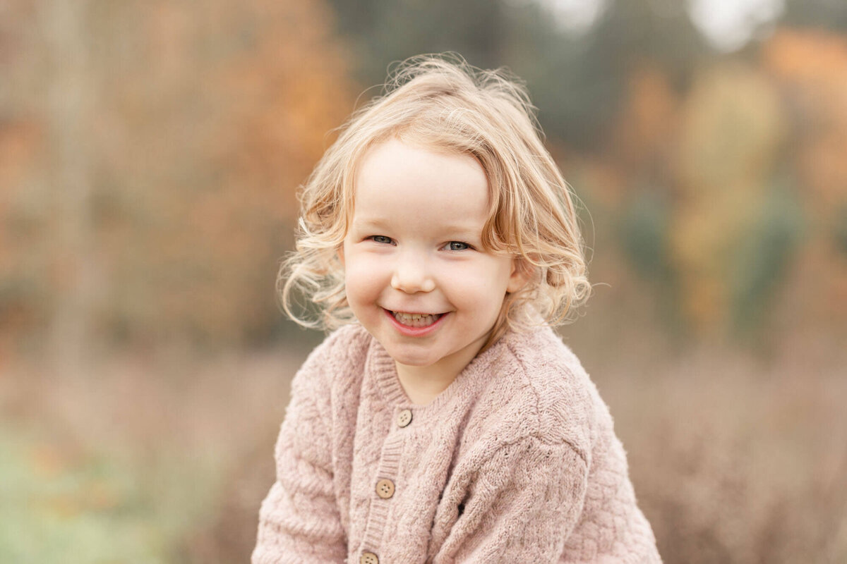Beautiful toddler girl with blonde curly hair wearing a pink sweater and smiling towards the camera. She is in a field of tall golden grasses and a tree line with fall colors that is blurred out behind her