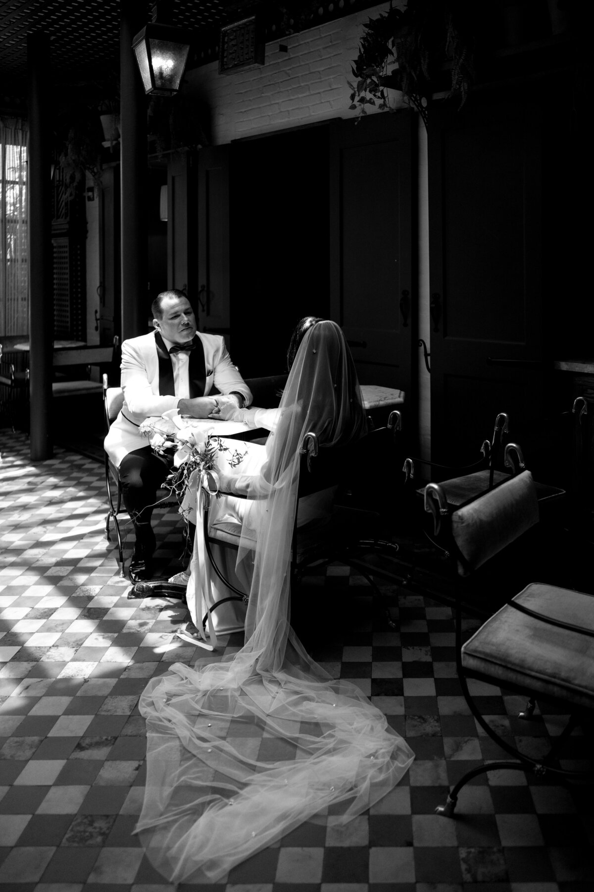 Bride and groom seated at a café table inside Hotel Chelsea, with the bride’s veil cascading to the floor, captured in black and white during Japna and Chris’s New York City elopement by wedding photographer Perry Hancock.